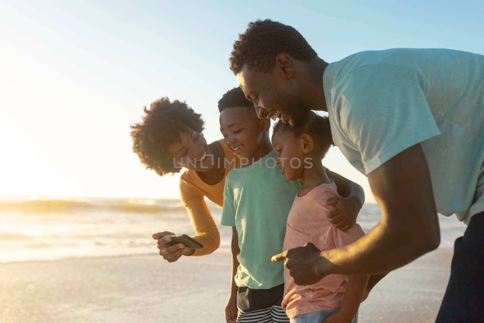 Happy african american woman sharing smartphone with family at beach during sunset. unaltered, family, lifestyle, togetherness, technology and holiday concept.