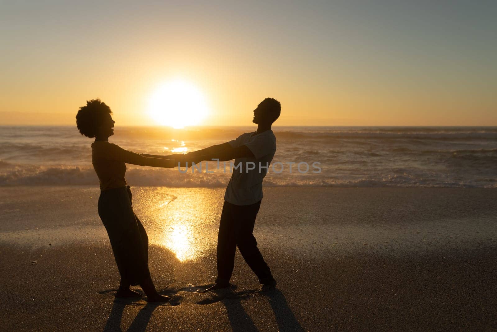 Full length side view of happy african american couple holding hands enjoying sunset at beach. unaltered, lifestyle, love and togetherness concept.