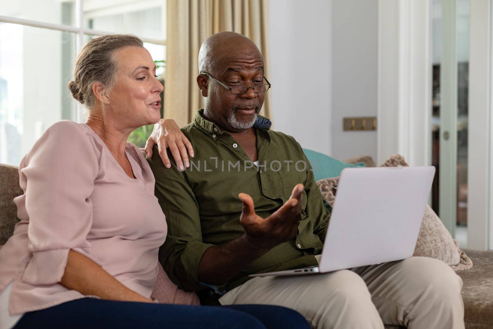 Senior multiracial couple discussing over laptop while sitting on sofa at home by Wavebreakmedia