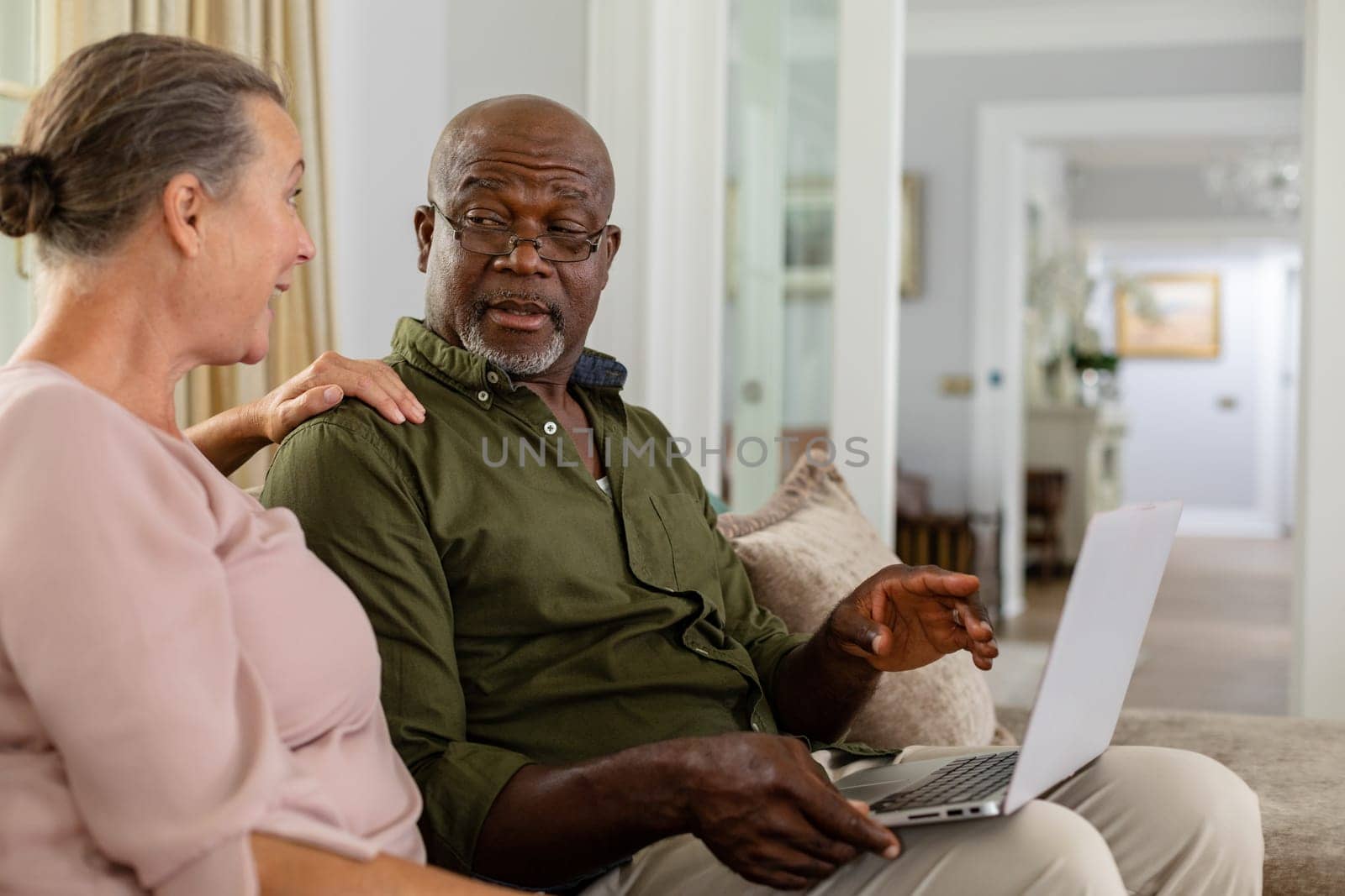 Senior multiracial couple with laptop discussing while sitting on sofa at home. unaltered, lifestyle, retirement, togetherness, leisure, wireless technology.