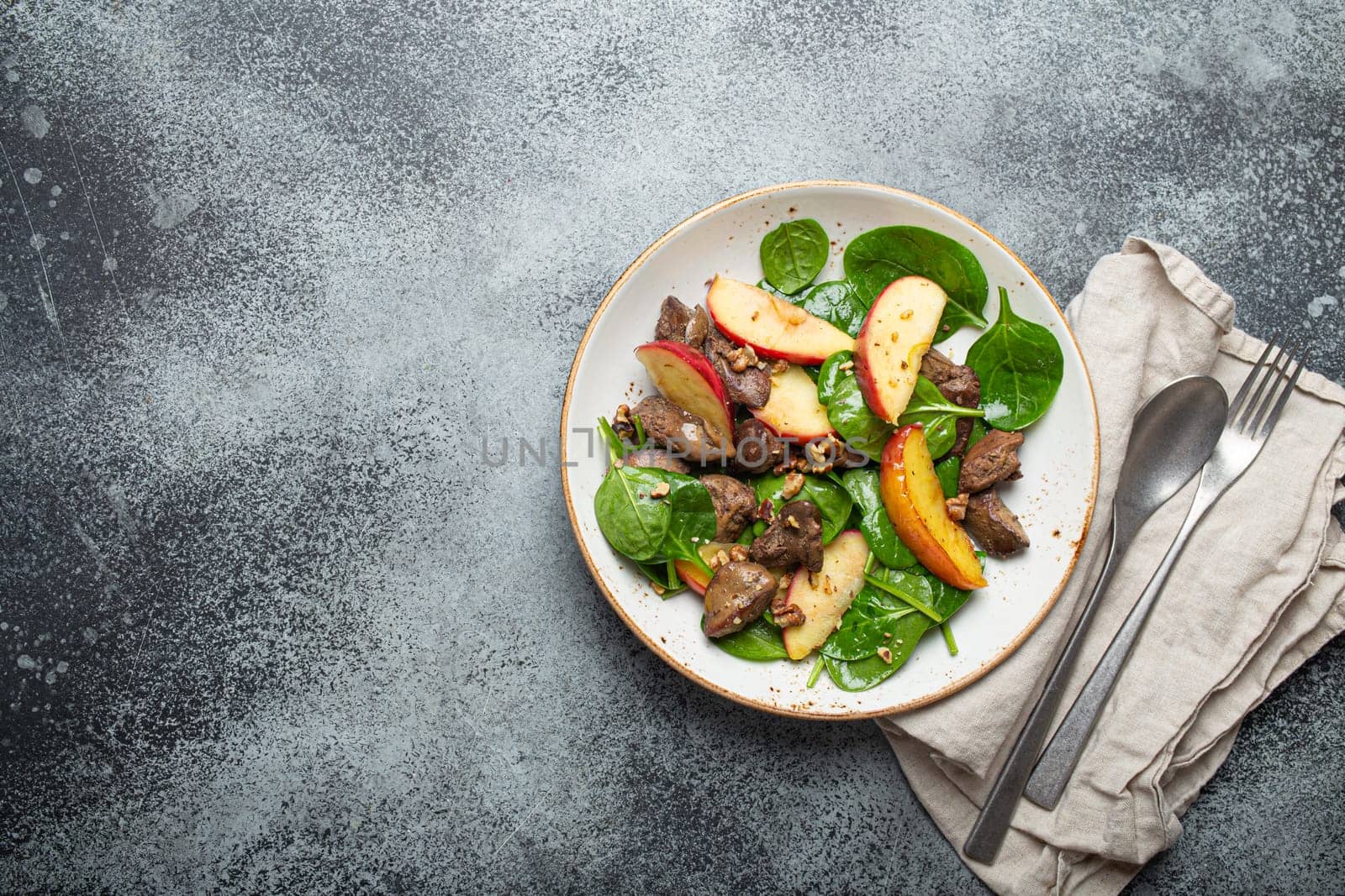 Healthy Salad with Iron Rich Ingredients Chicken Liver, Apples, Fresh Spinach and Walnuts on White Ceramic Plate, Grey Rustic Stone Background Top View Copy Space.