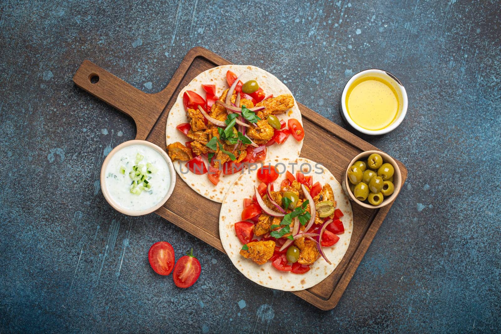 Cooking Traditional Greek Dish Gyros: Pita bread with vegetables, meat, herbs, olives on rustic wooden cutting board with Tzatziki sauce, olive oil top view on dark blue stone background.