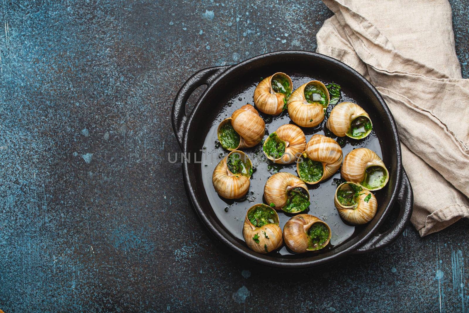 Escargots de Bourgogne Snails with Garlic Butter and Parsley in black cast iron pan on rustic stone background top view, traditional French Delicacy, space for text.