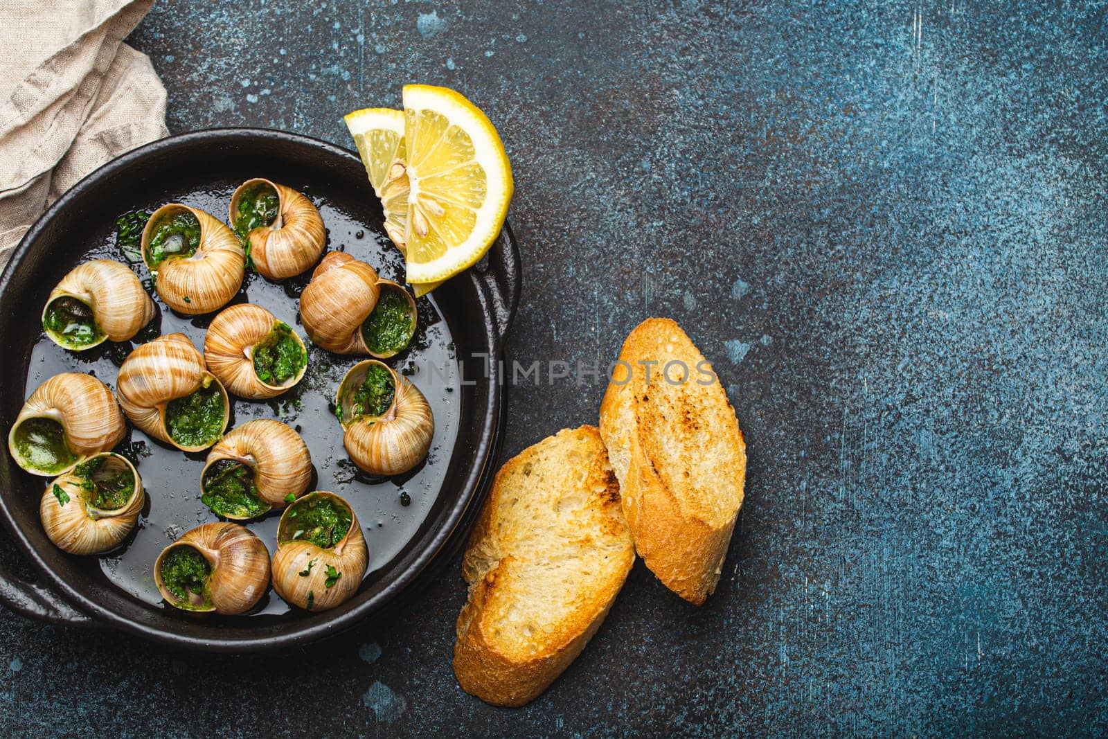 Escargots de Bourgogne Snails with Garlic Butter and Parsley in black cast iron pan with Lemon and Toasted Baguette Slices on rustic stone background top view, traditional French Delicacy, copy space.