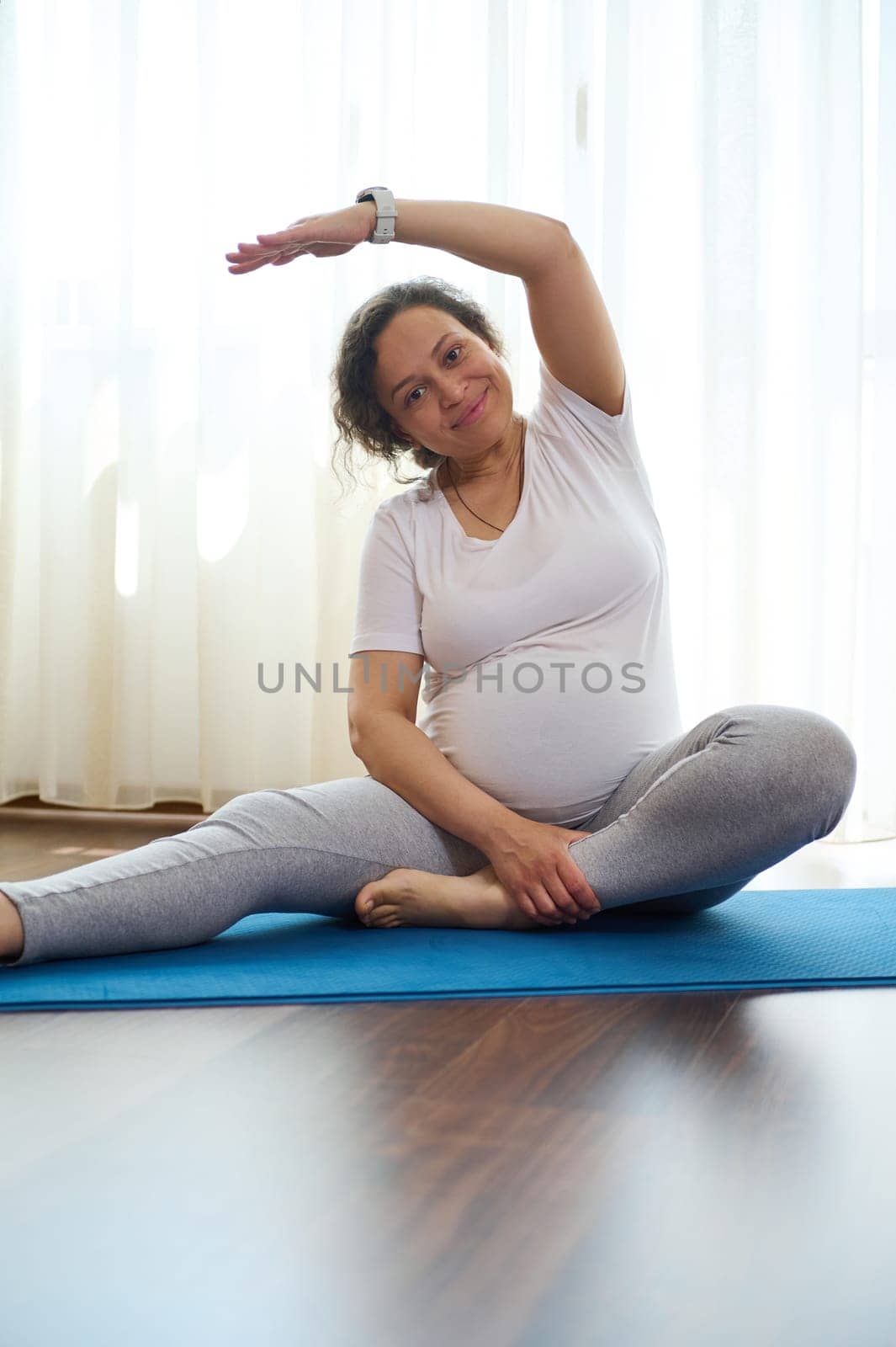 Smiling pregnant woman stretching her body while exercising on yoga mat at home. The concept of healthy active pregnancy and happy maternity leave. Prenatal fitness. Pregnancy. Maternity lifestyle