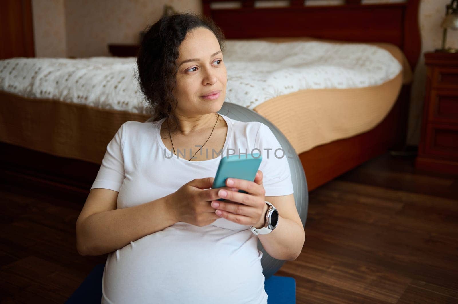 Multi-ethnic thoughtful pretty gravid female, pregnant woman, expectant mother holding mobile phone, checking new application, shopping online, dreamily looking aside, sitting on yoga mat at home
