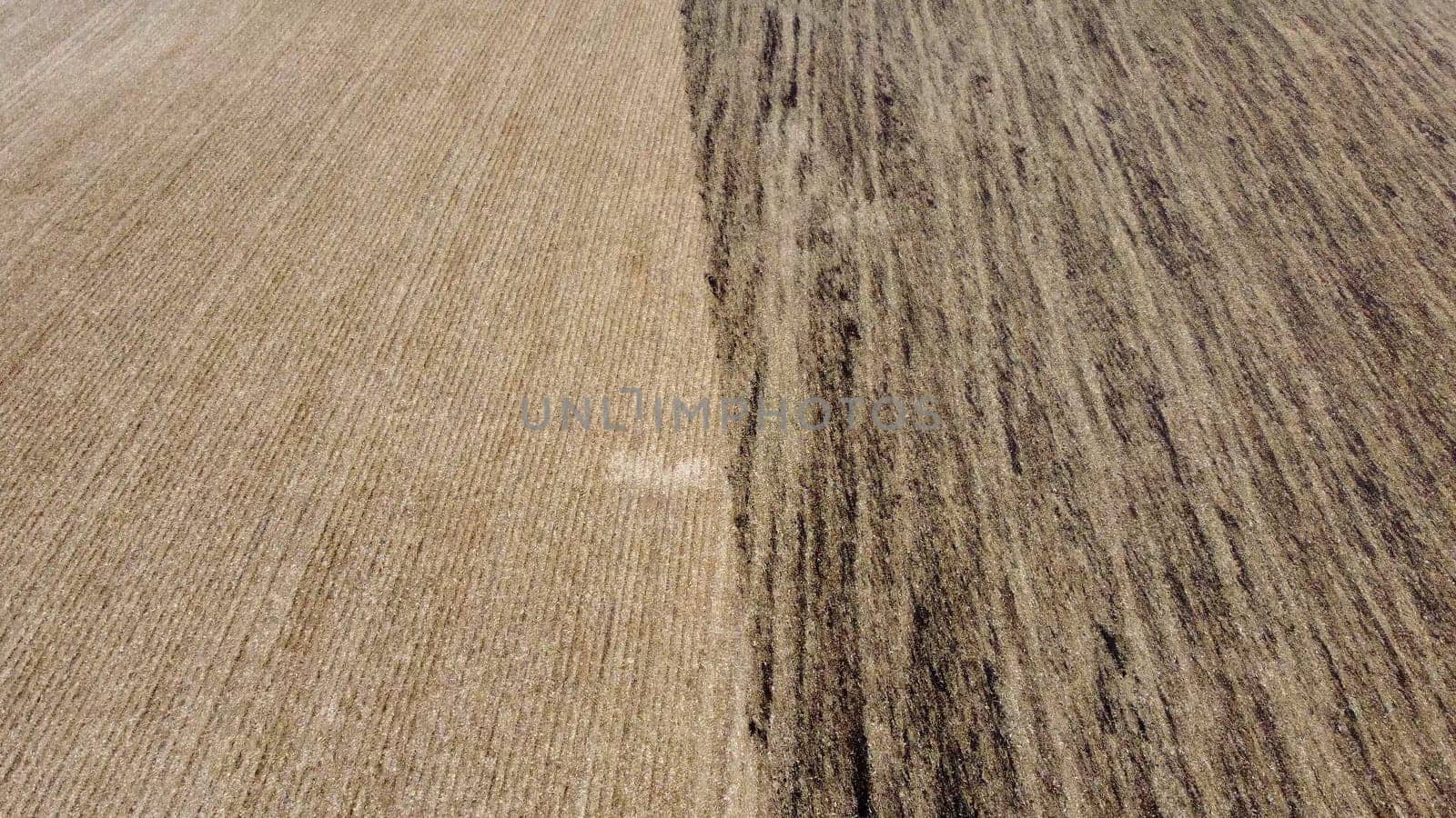 Half of large field after wheat harvest, half of field plowed, motorway with moving car and grain elevator. Flying over field with dry straw from wheat and brown dug up earth. Agricultural background.