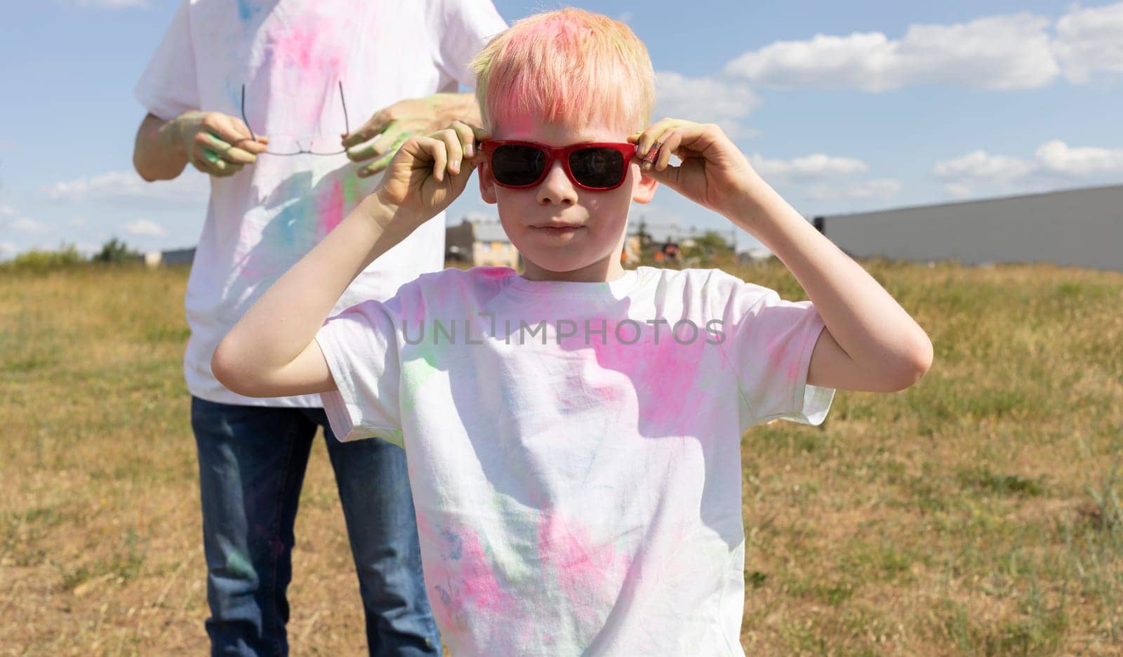 Closeup Little Boy With Colorful Paint, Powder On Cloth And Face Posing On Holi Color Festival Or Birthday In Meadow. Father Is On Background, Sunny Day. Horizontal Plane. High quality photo