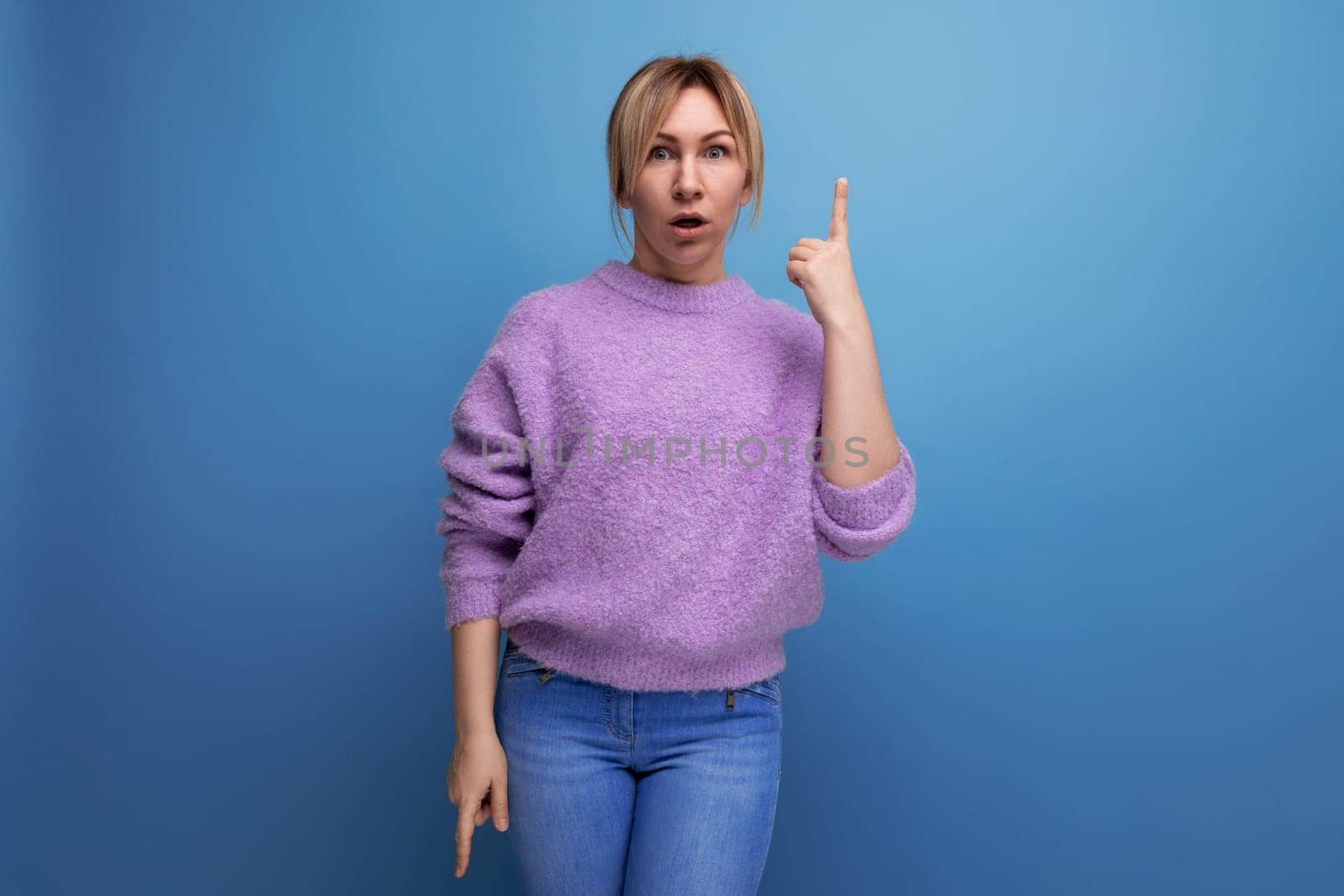 surprised pleasant blond young woman in purple sweater shows hand up on blue background with copy space.