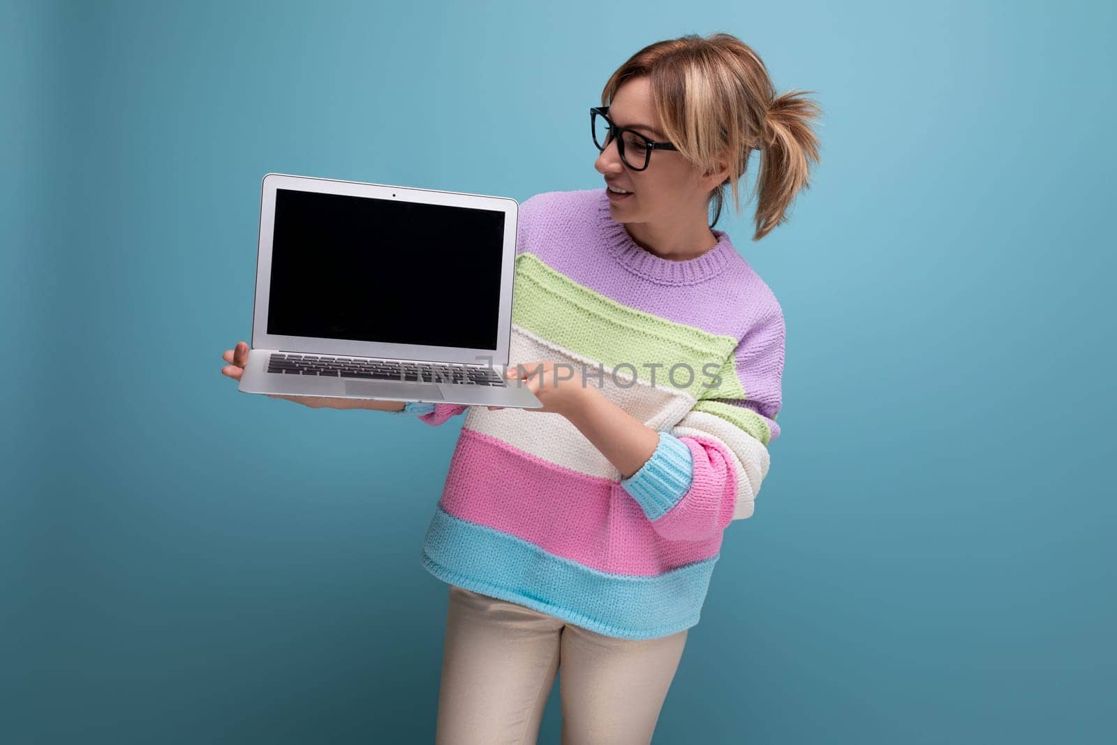 Attractive successful blond female freelancer in a striped sweater demonstrates a mockup of a laptop on a blue background with copy space.