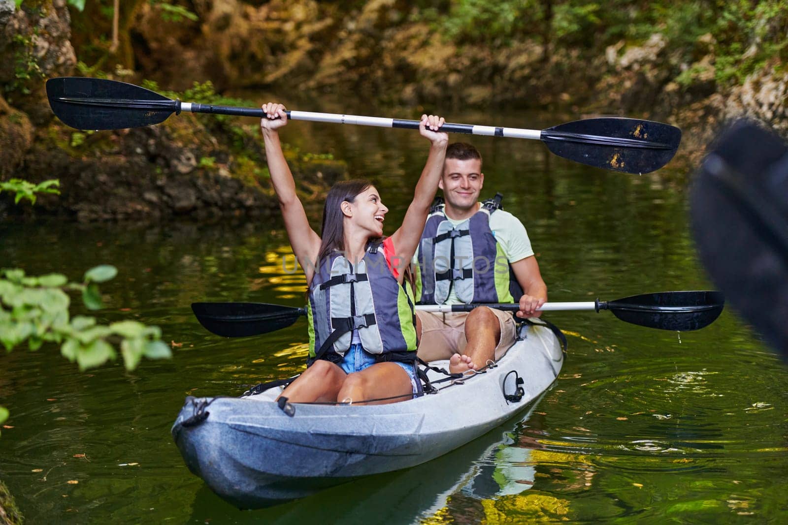 A young couple enjoying an idyllic kayak ride in the middle of a beautiful river surrounded by forest greenery by dotshock