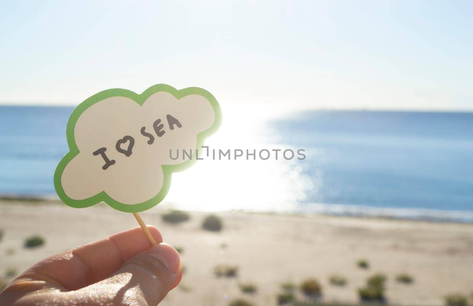 Stick with paper speech bubble with words I love sea on background blue sea sky sandy beach on sunny summer day. Text-balloons with text from letters. Concept symbol sign vacation travel tourism rest