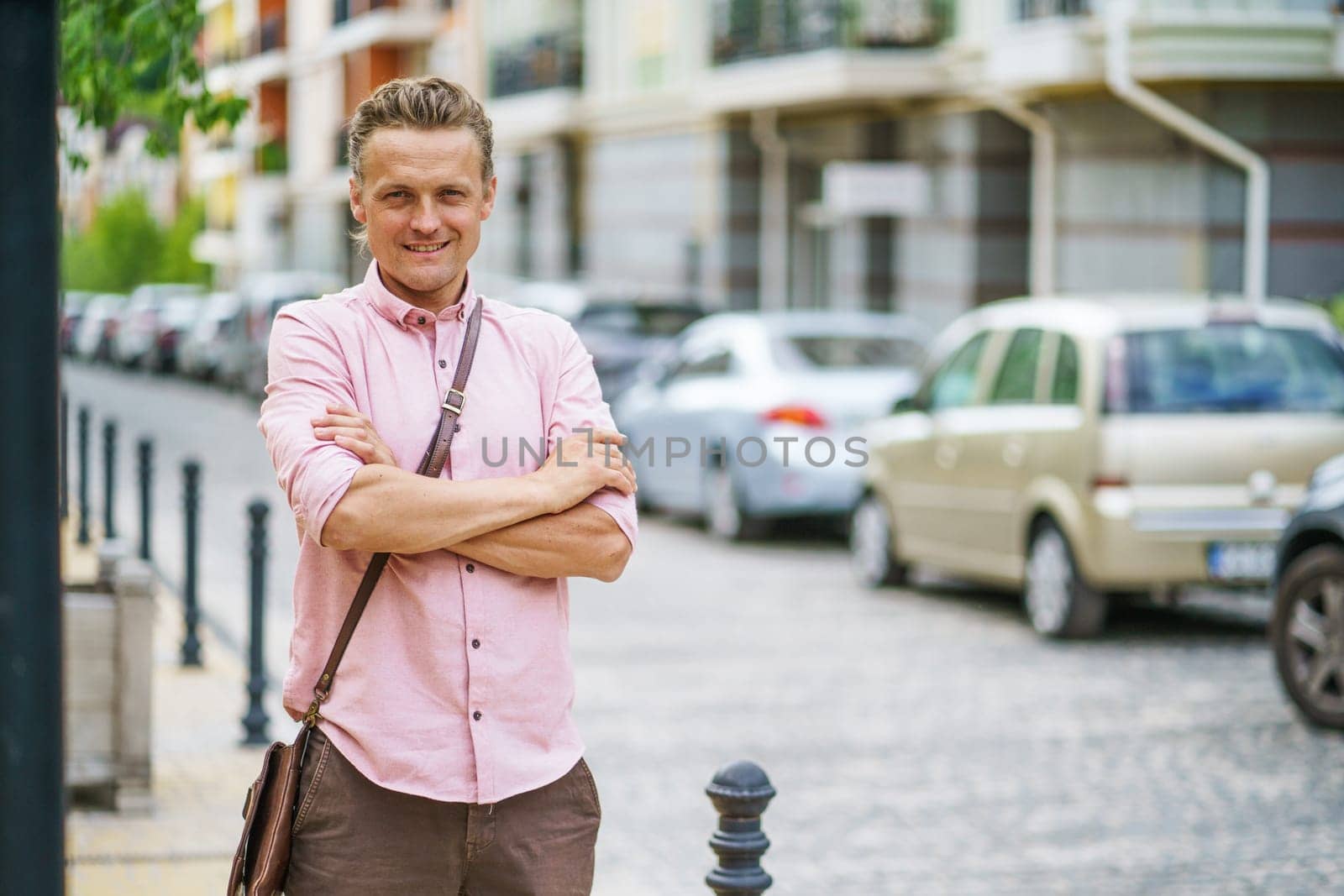 Blond, handsome Caucasian individual confidently standing on street in city of Europe, with crossed hands. Person's stylish appearance and confident stance add to their overall charm. . High quality photo