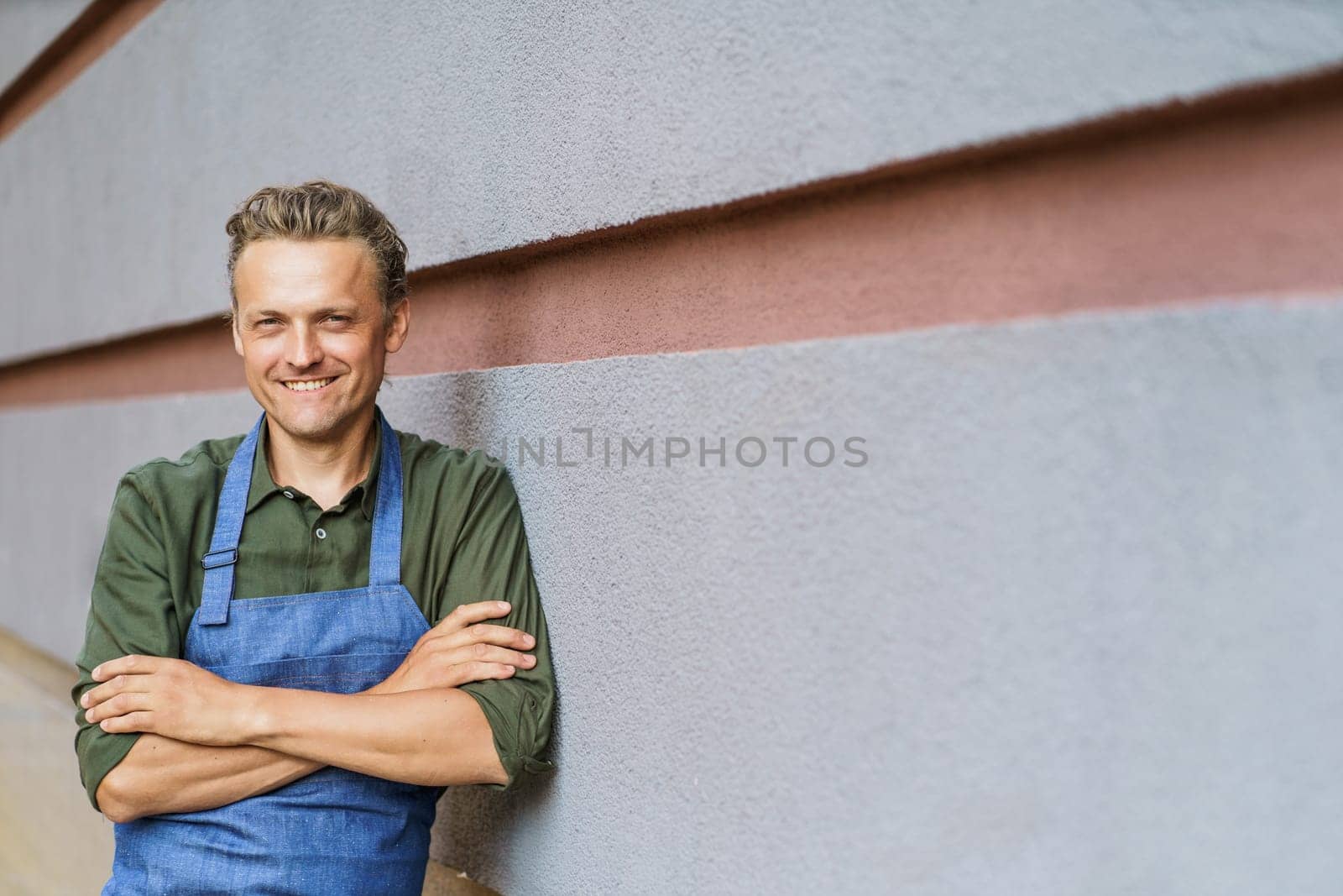 Mid-aged waiter in apron, smiling while standing near wall with crossed hands. With friendly and welcoming smile, waiter embodies essence of customer service in hospitality industry. . High quality photo