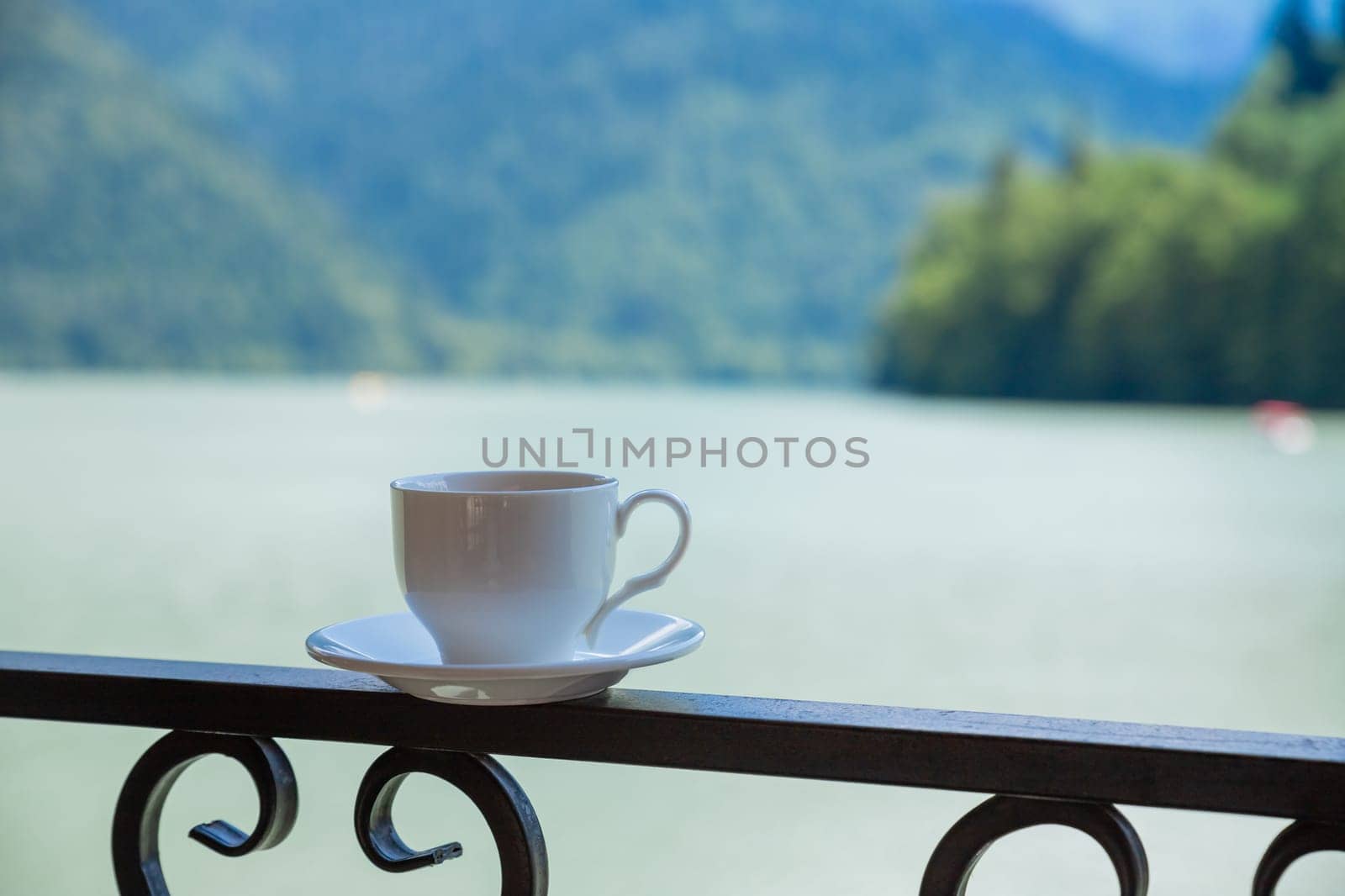 Cup of hot coffee on terrace with beautiful view of Alpine mountain landscape at background with small lake downhill in valley.