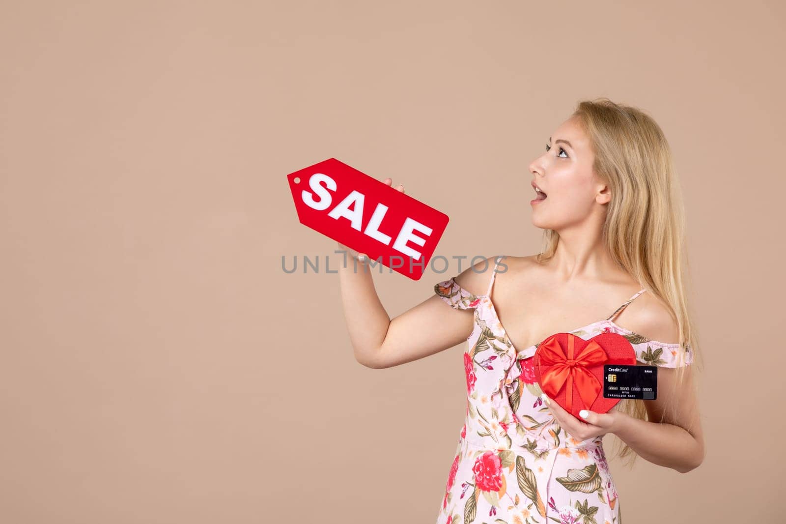 front view young female holding red present sale nameplate and bank card on brown background money march horizontal feminine equality sensual woman by Kamran