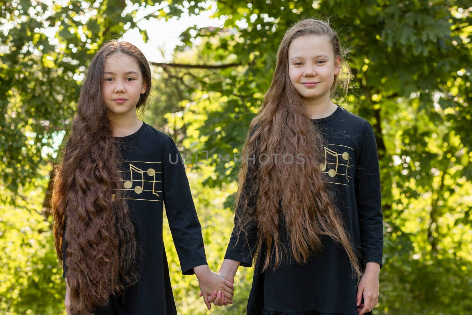 Portrait Smiling Two Sisters With Extra Long Hair Holds Hands In Sunny Day, Girls Wear Black Dresses. Pretty Siblings, Friendship. Green Trees On Background. Horizontal Plane. High Quality Photo