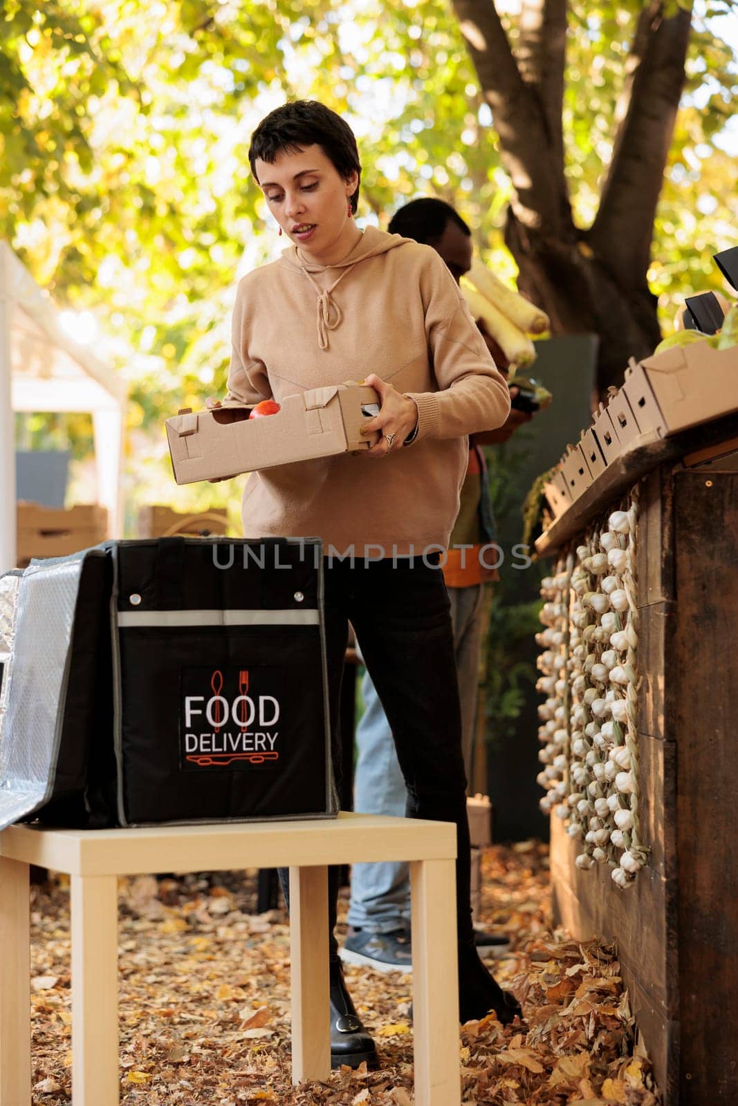 Young adult with backpack delivering organic produce in box, preparing to take natural food order to customers. Woman delivery courier picking up order from local market stand.