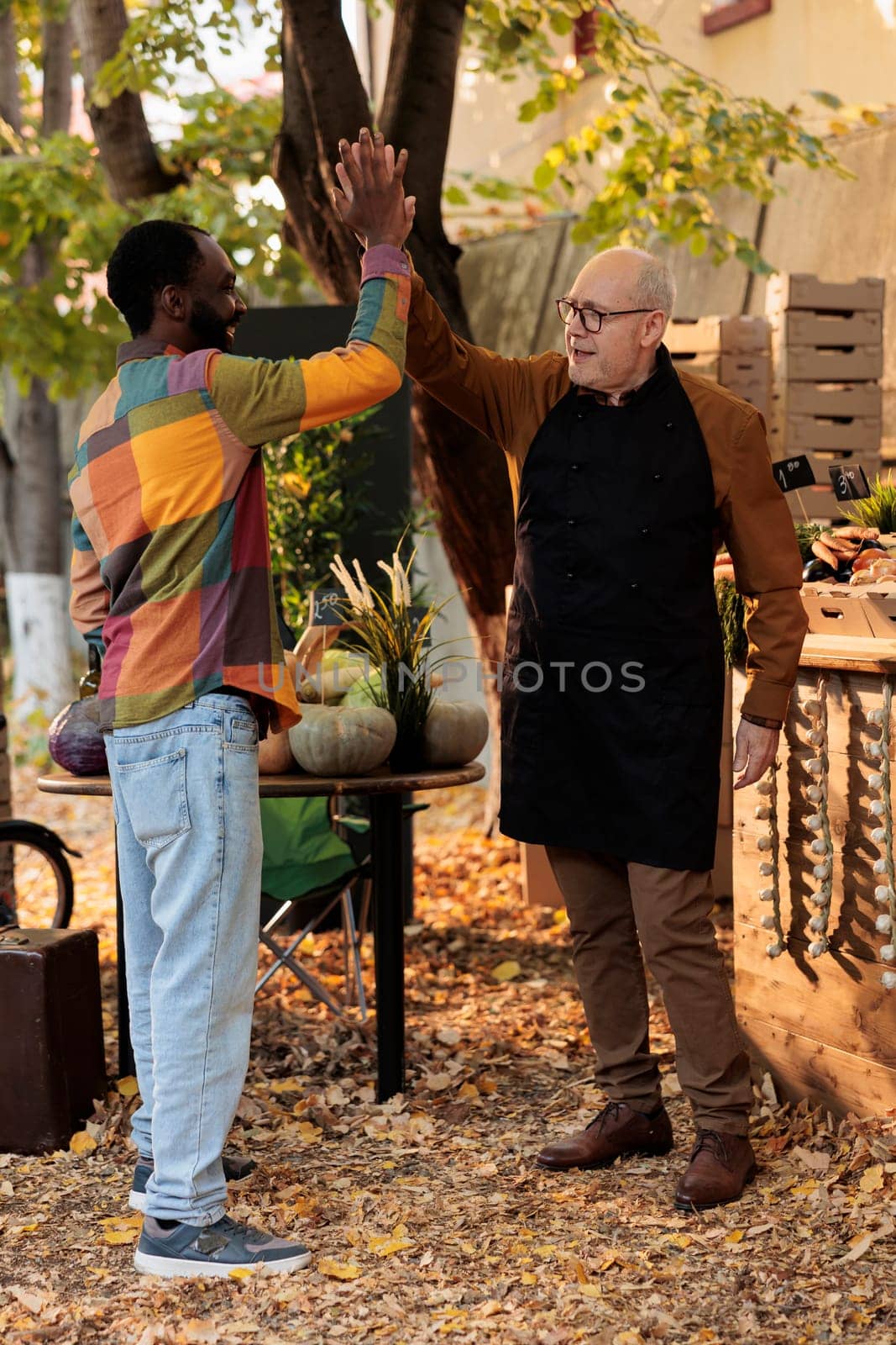 Positive small business owner greeting man with highfive, enjoying visit from loyal customer at local farmers market stand. Smiling happy people having fun at food fair, organic fresh produce.