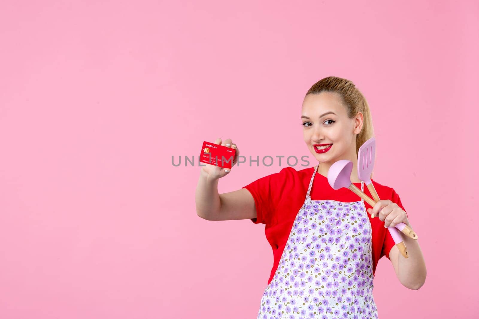 front view young housewife in cape holding spoons and bank card on pink background duty money worker horizontal wife uniform job cutlery by Kamran