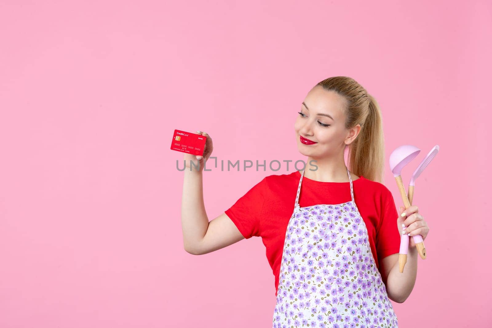 front view young housewife in cape holding spoons and bank card on pink background occupation cutlery horizontal job wife duty profession money uniform by Kamran