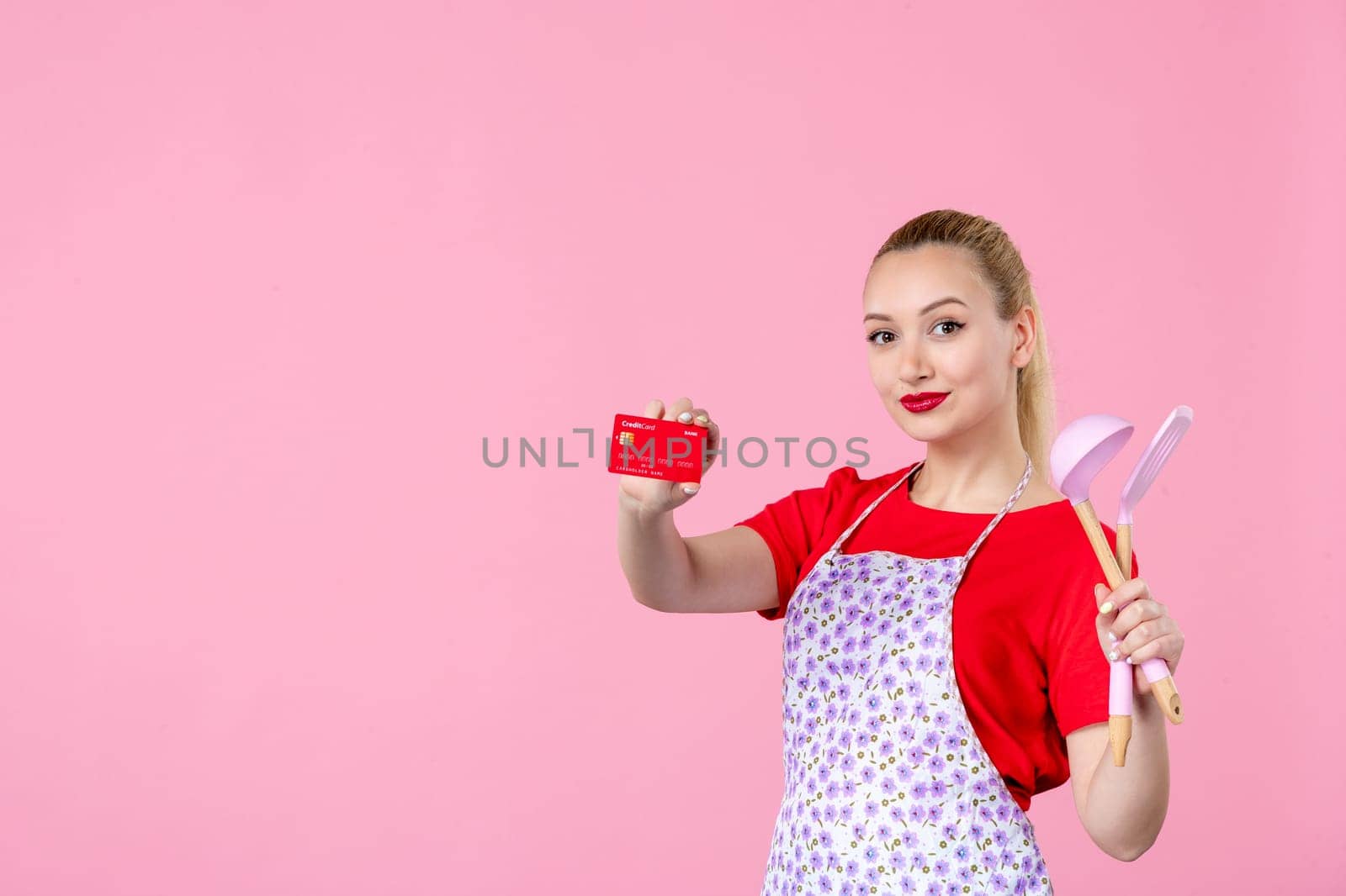 front view young housewife in cape holding spoons and bank card on pink background occupation duty money horizontal wife profession uniform job cutlery