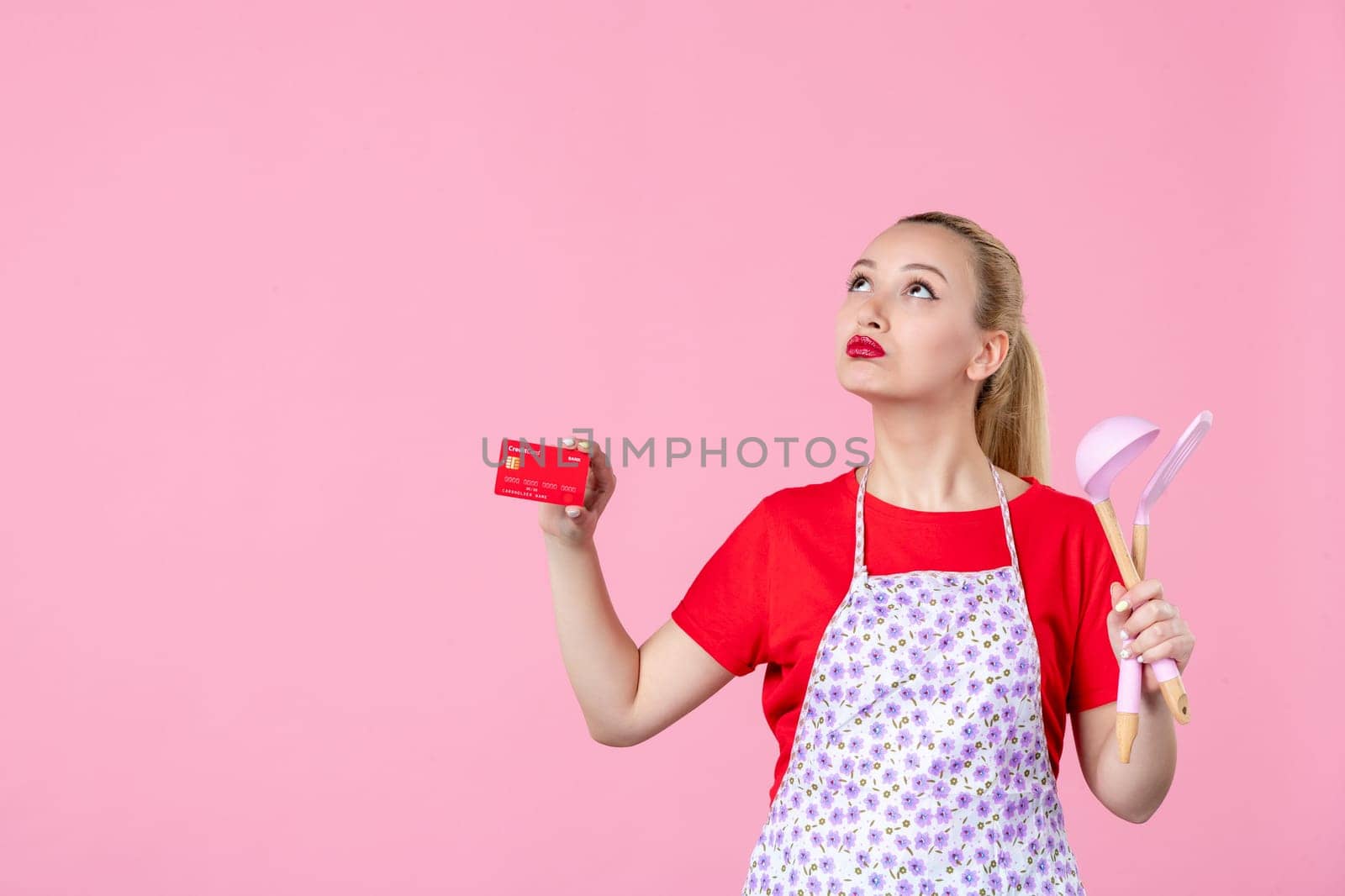 front view young housewife in cape holding spoons and bank card on pink background occupation duty money worker horizontal profession uniform job cutlery