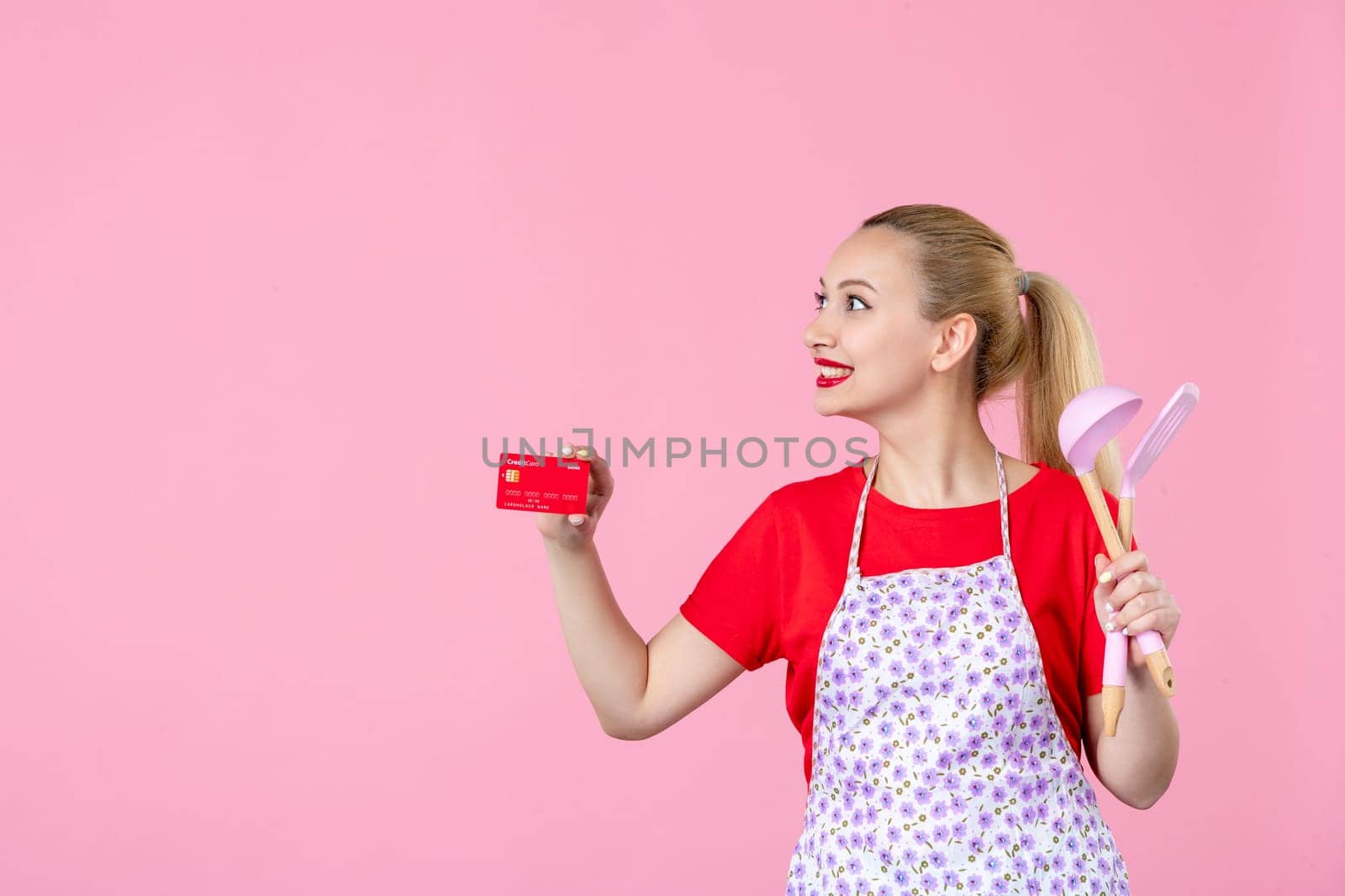 front view young housewife in cape holding spoons and bank card on pink background occupation duty worker cutlery horizontal wife profession money uniform by Kamran