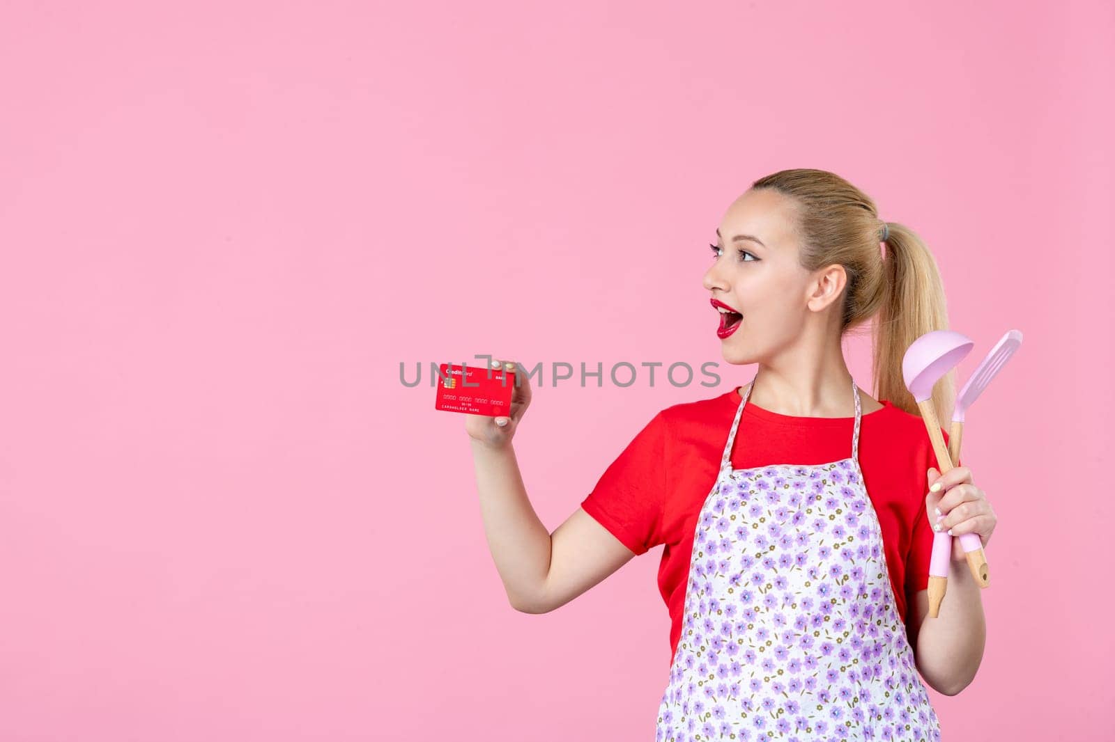 front view young housewife in cape holding spoons and bank card on pink background occupation duty worker cutlery horizontal wife profession uniform job