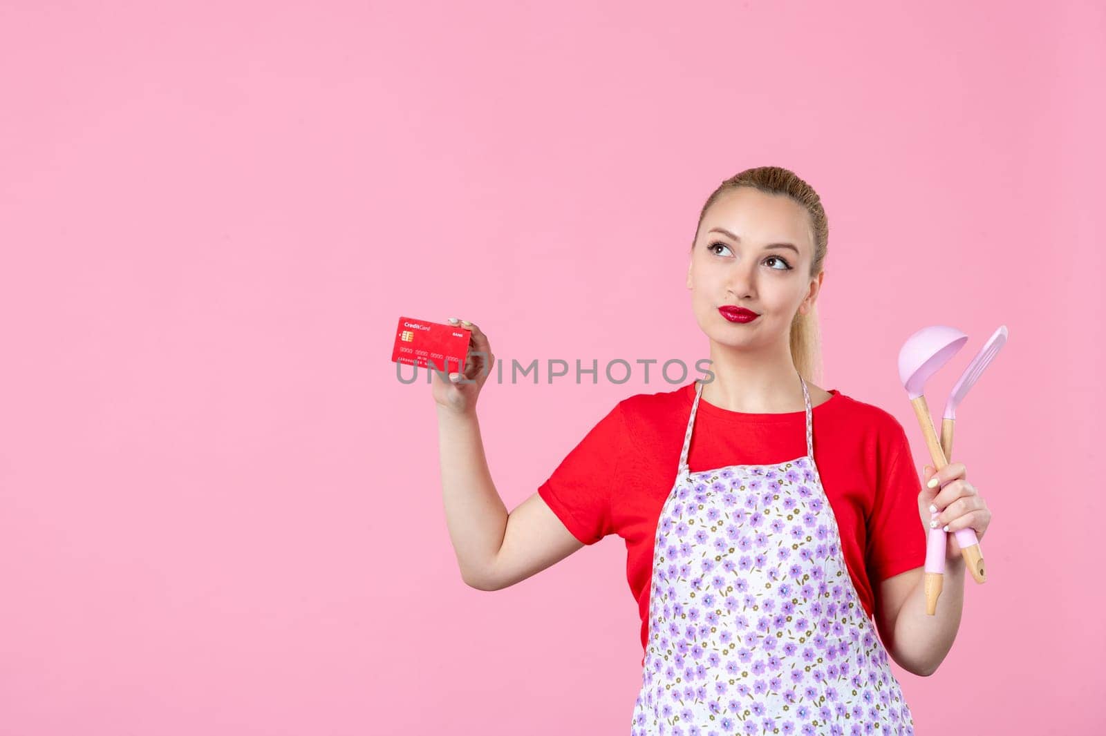 front view young housewife in cape holding spoons and bank card on pink background occupation worker cutlery job wife duty profession money uniform by Kamran