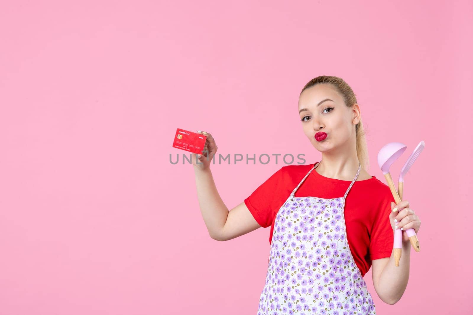front view young housewife in cape holding spoons and bank card on pink background occupation worker horizontal job wife duty profession money uniform