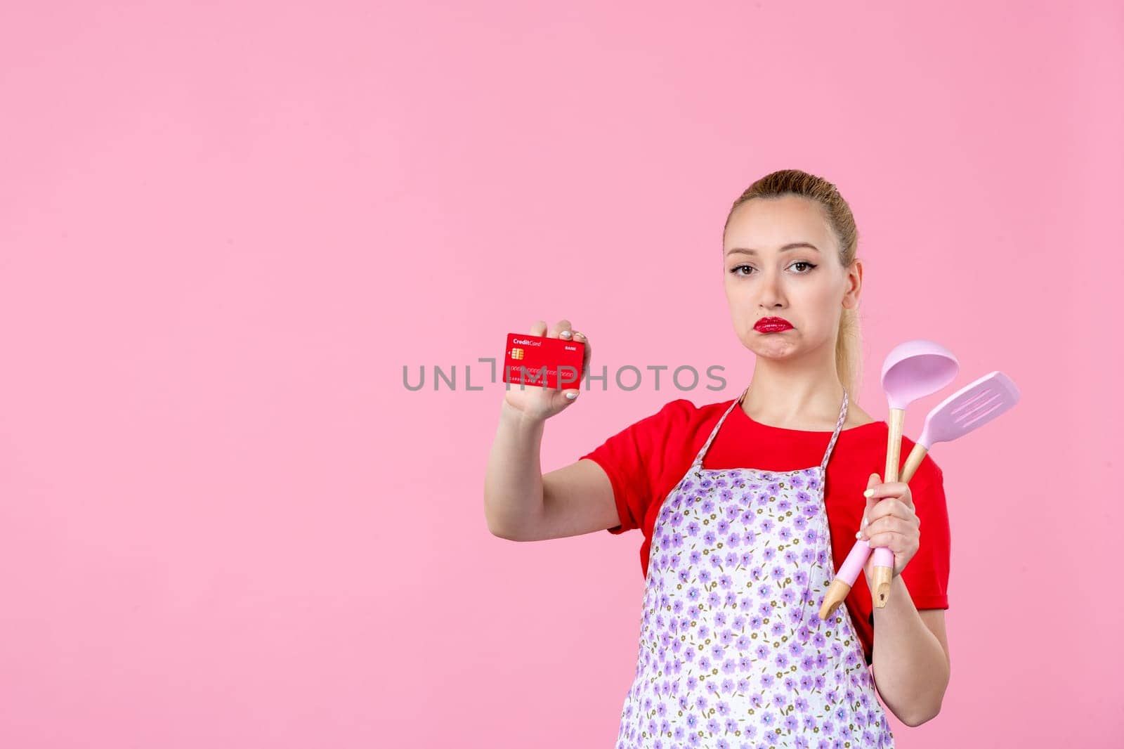 front view young housewife in cape holding spoons and bank card on pink background profession duty money horizontal wife uniform job cutlery worker