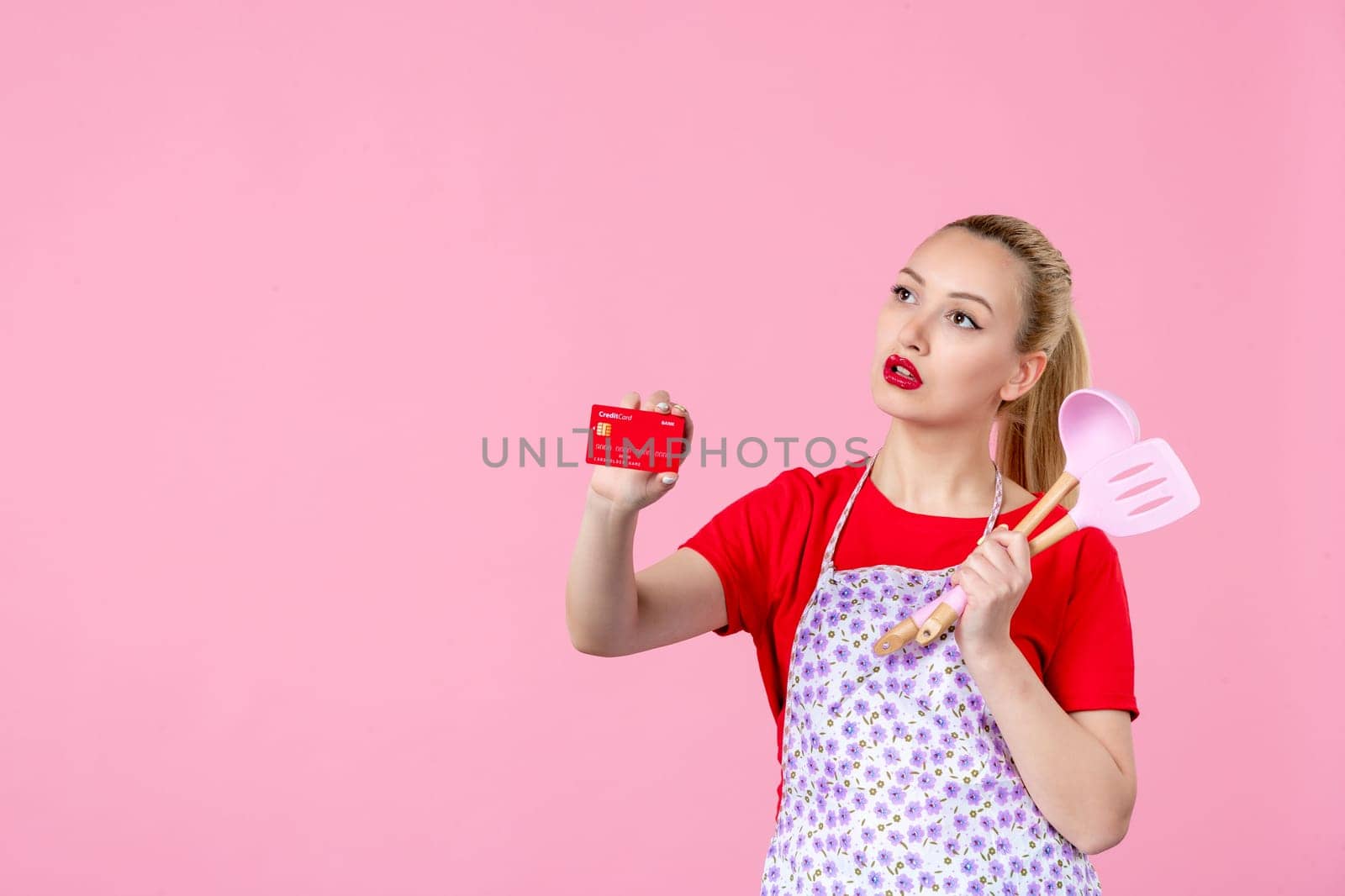 front view young housewife in cape holding spoons and bank card on pink background profession occupation duty money wife uniform cutlery worker horizontal