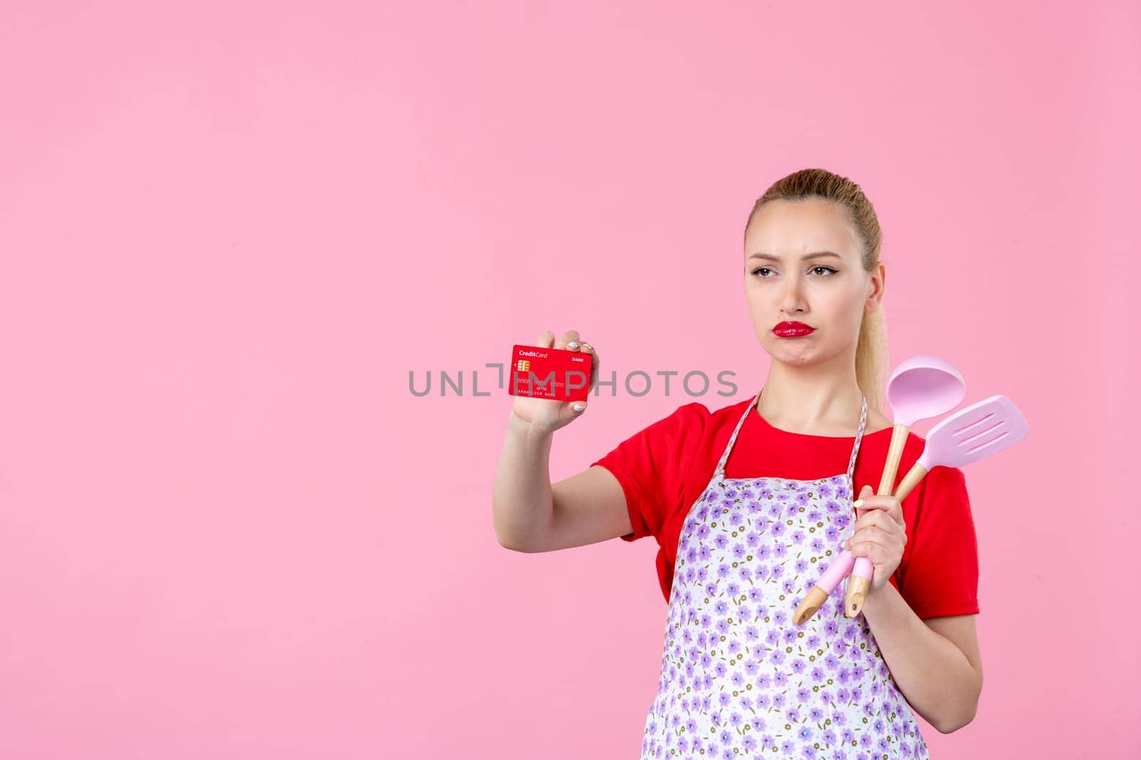 front view young housewife in cape holding spoons and bank card on pink background profession occupation duty money wife uniform job cutlery worker horizontal