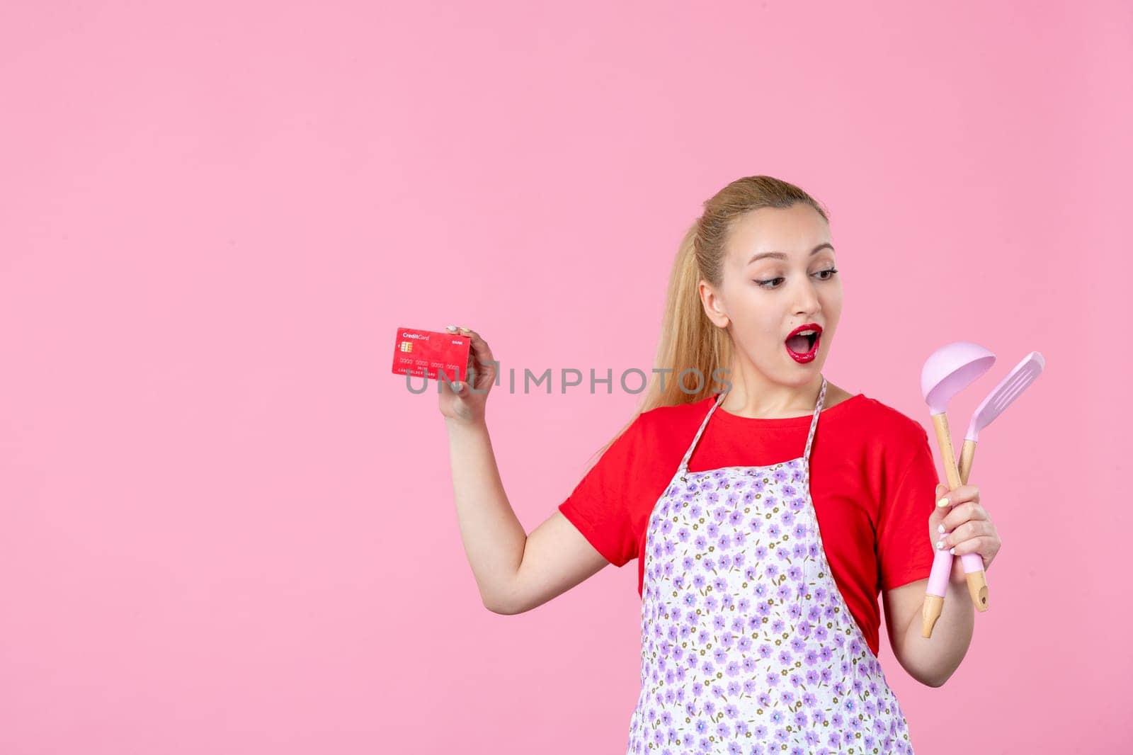 front view young housewife in cape holding spoons and bank card on pink background worker cutlery uniform horizontal job wife duty profession by Kamran