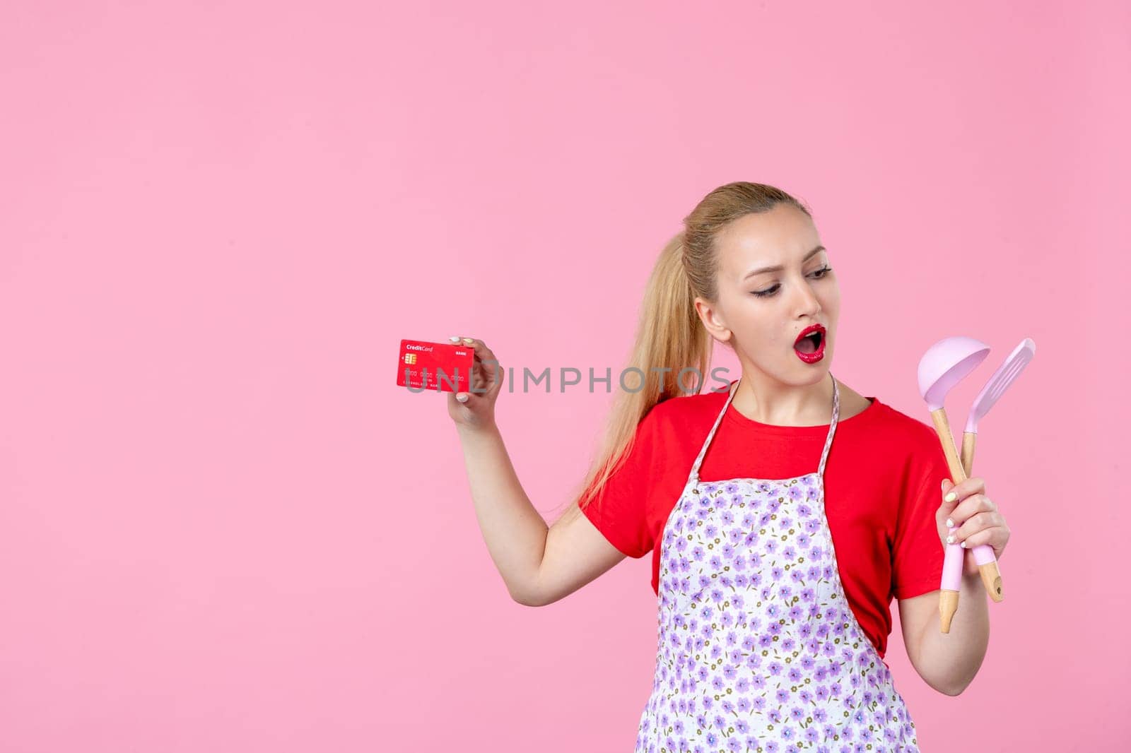 front view young housewife in cape holding spoons and bank card on pink background worker cutlery uniform horizontal occupation job wife duty