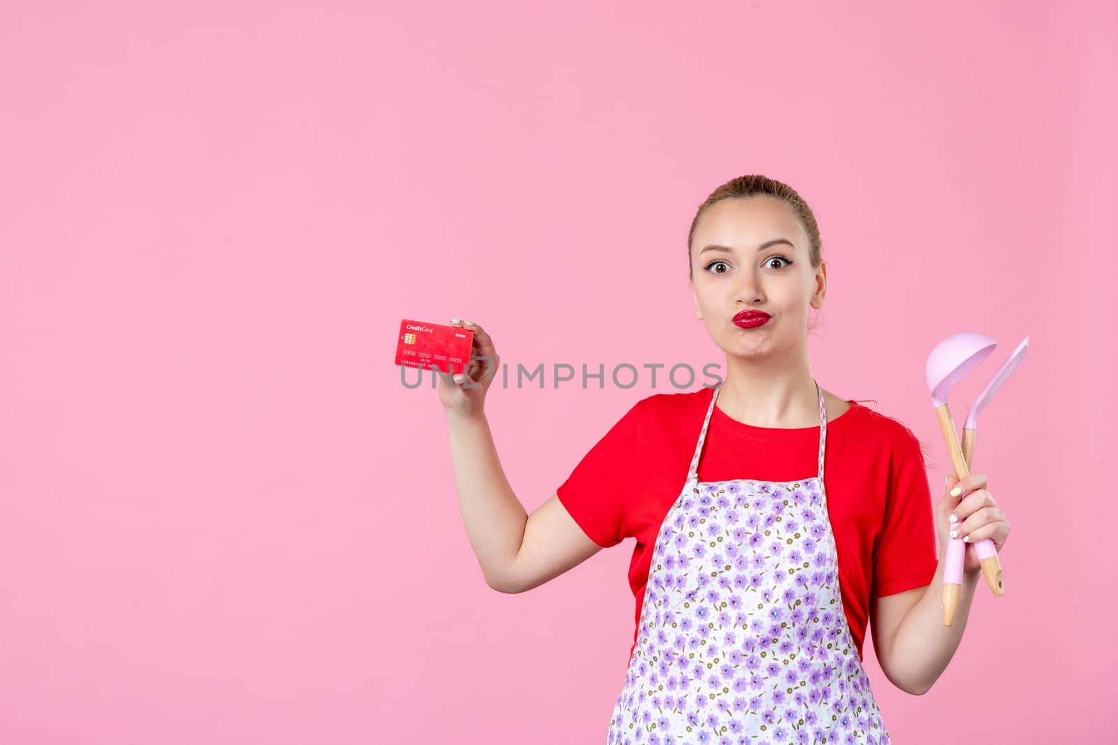front view young housewife in cape holding spoons and bank card on pink background worker cutlery uniform horizontal occupation job wife duty profession money