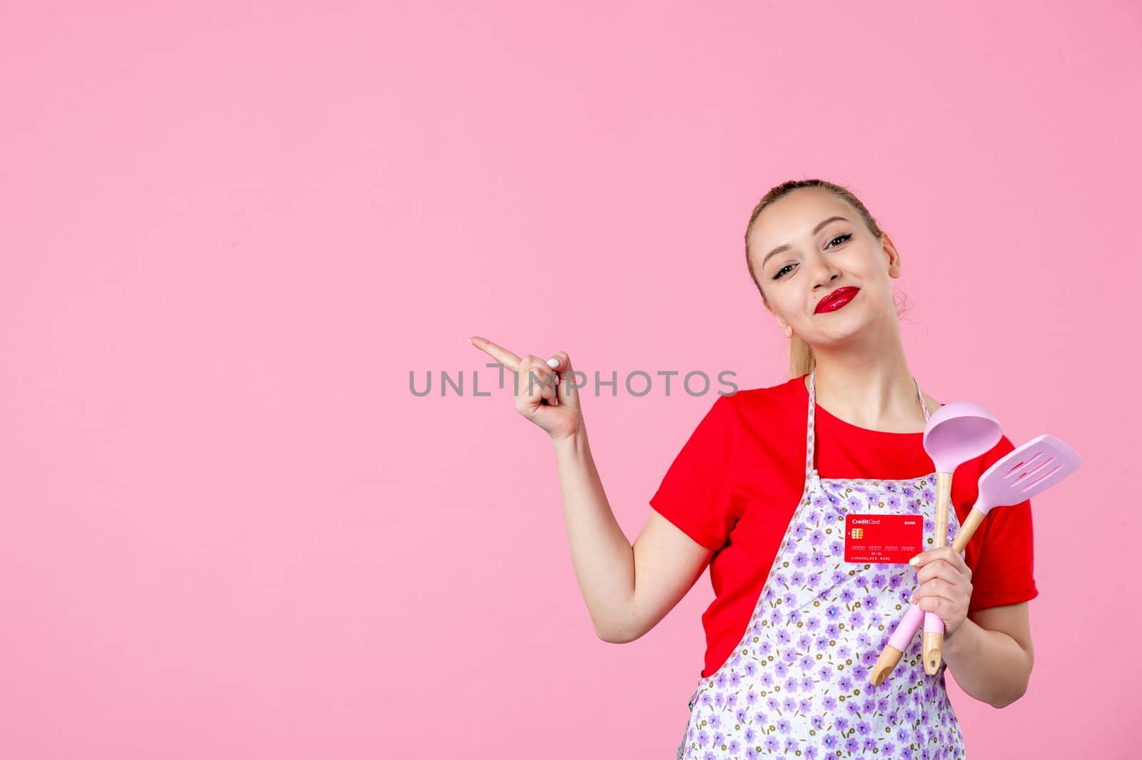 front view young housewife in cape holding spoons and red bank card on pink background occupation profession job horizontal wife duty worker uniform