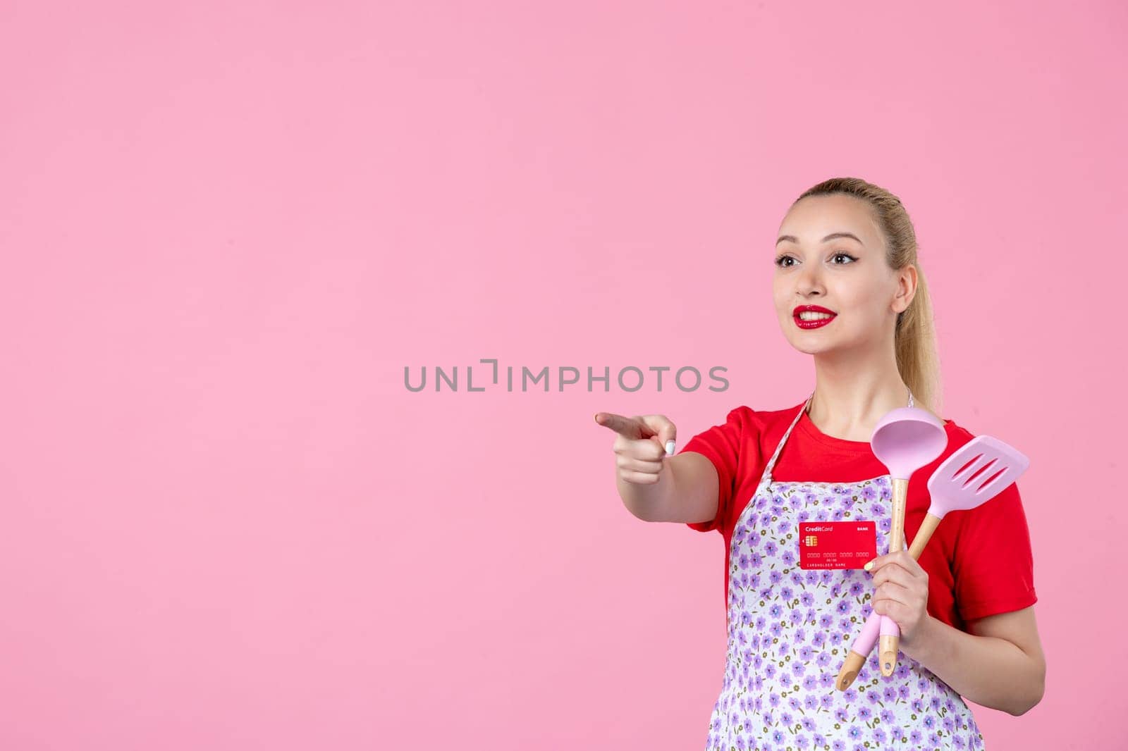 front view young housewife in cape holding spoons and red bank card on pink background occupation profession job horizontal wife money duty uniform by Kamran