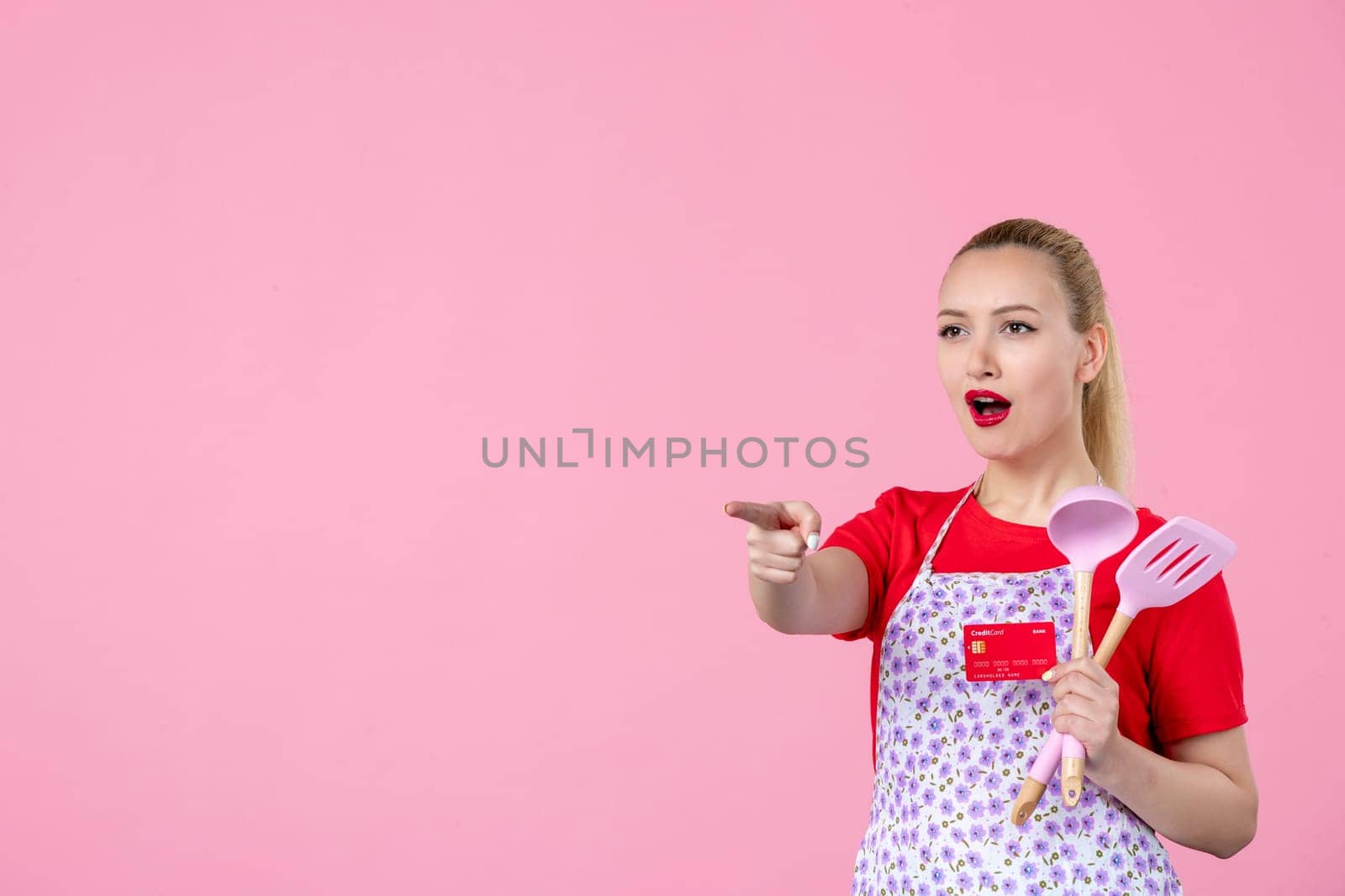 front view young housewife in cape holding spoons and red bank card on pink background occupation profession job horizontal wife money duty worker by Kamran