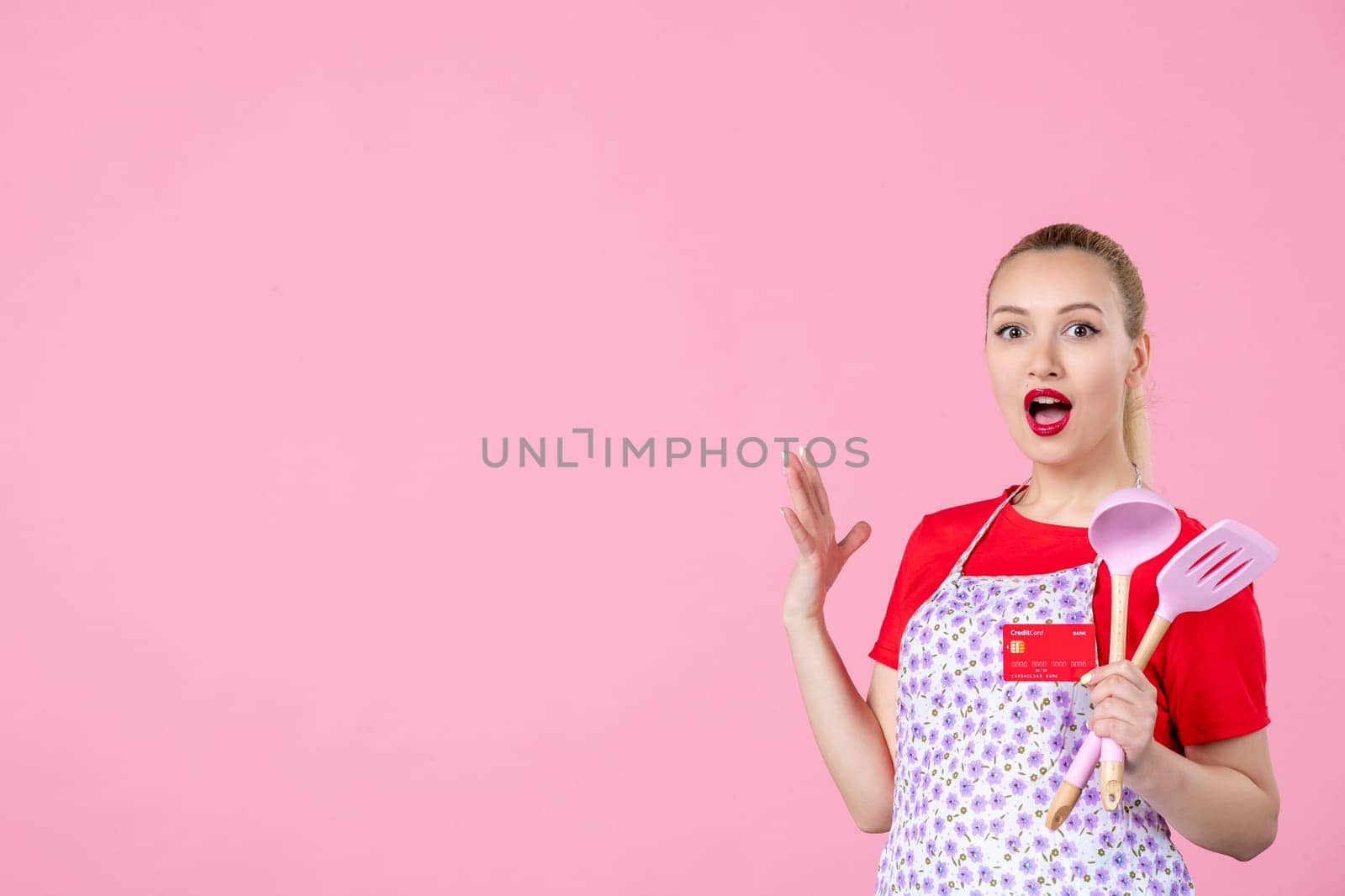 front view young housewife in cape holding spoons and red bank card on pink background occupation profession job horizontal wife money duty worker uniform by Kamran