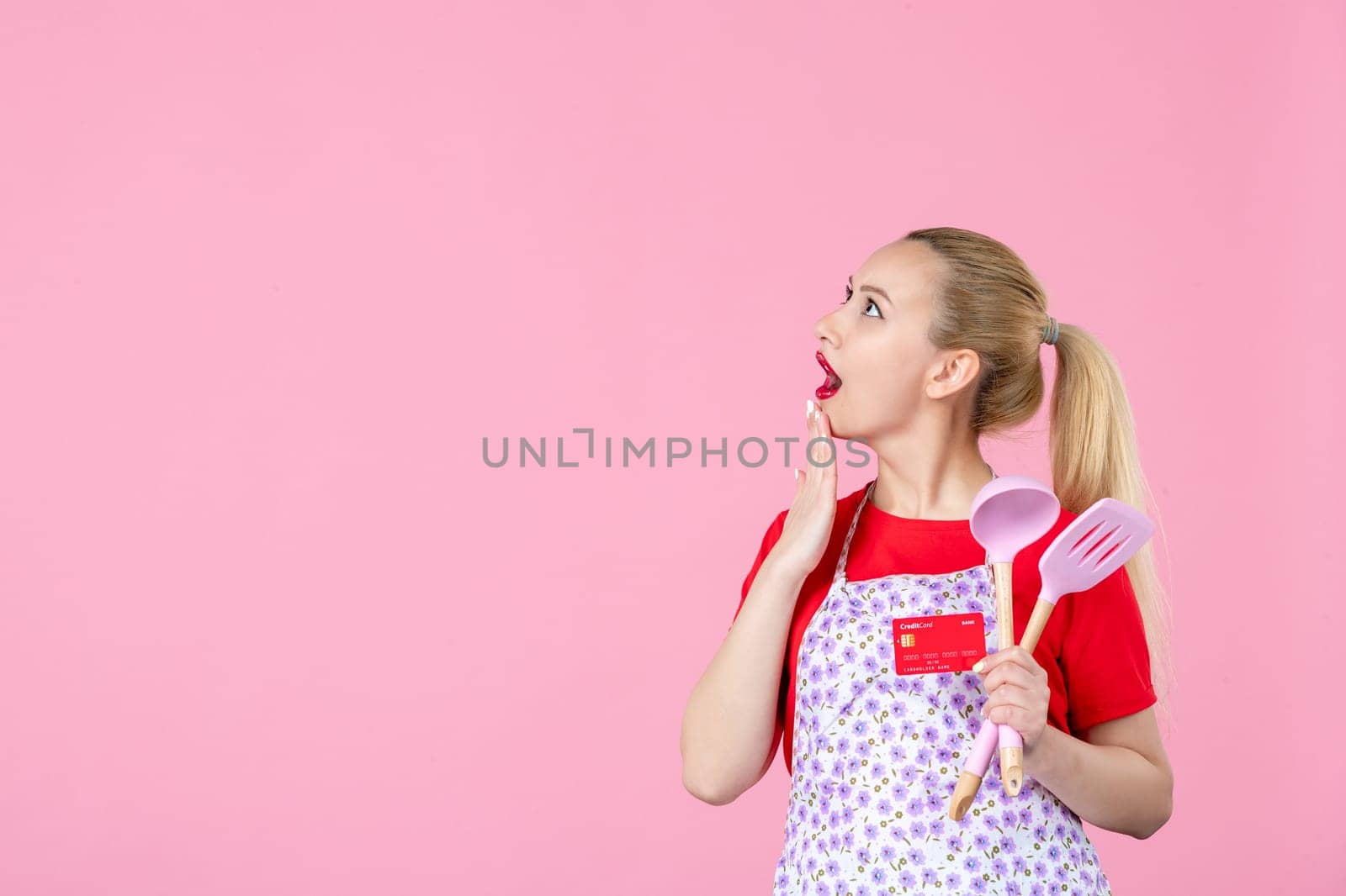 front view young housewife in cape holding spoons and red bank card on pink background occupation uniform profession job horizontal money duty by Kamran