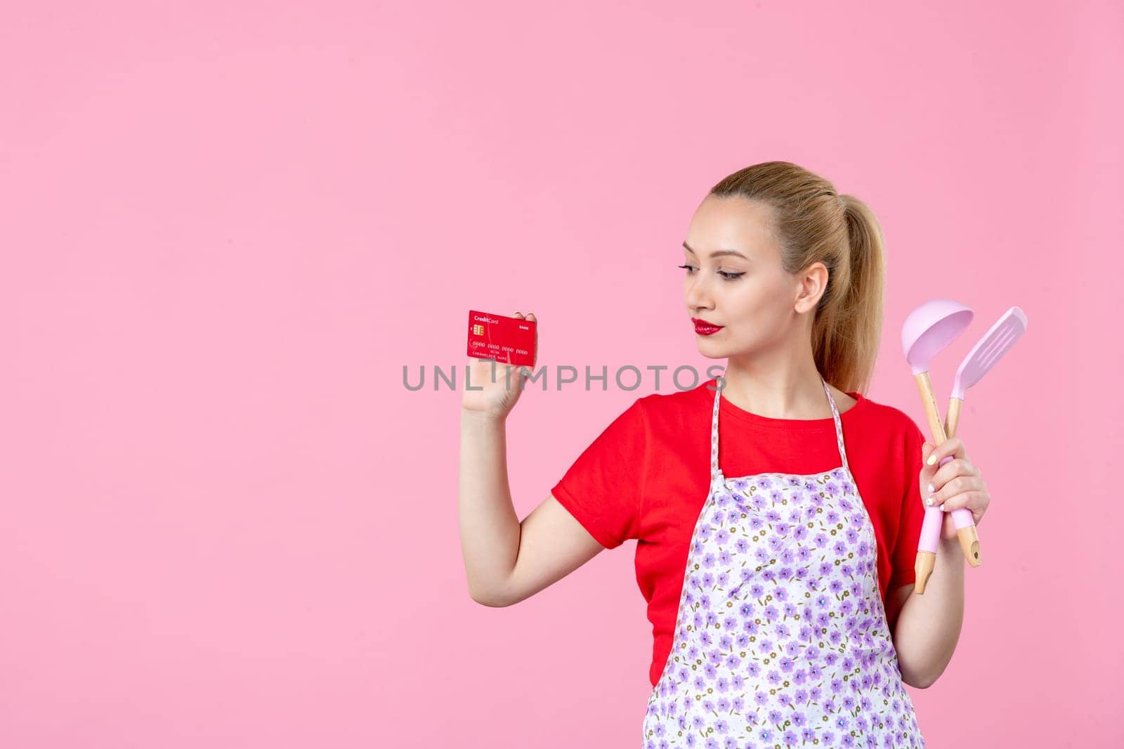 front view young housewife in cape holding spoons and red bank card on pink background occupation uniform profession job horizontal wife money duty
