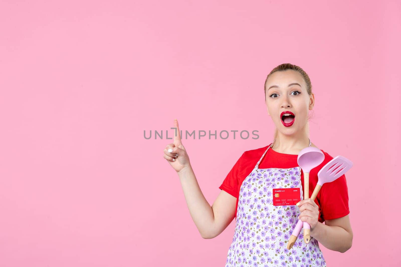 front view young housewife in cape holding spoons and red bank card on pink background occupation uniform profession job horizontal wife money duty worker