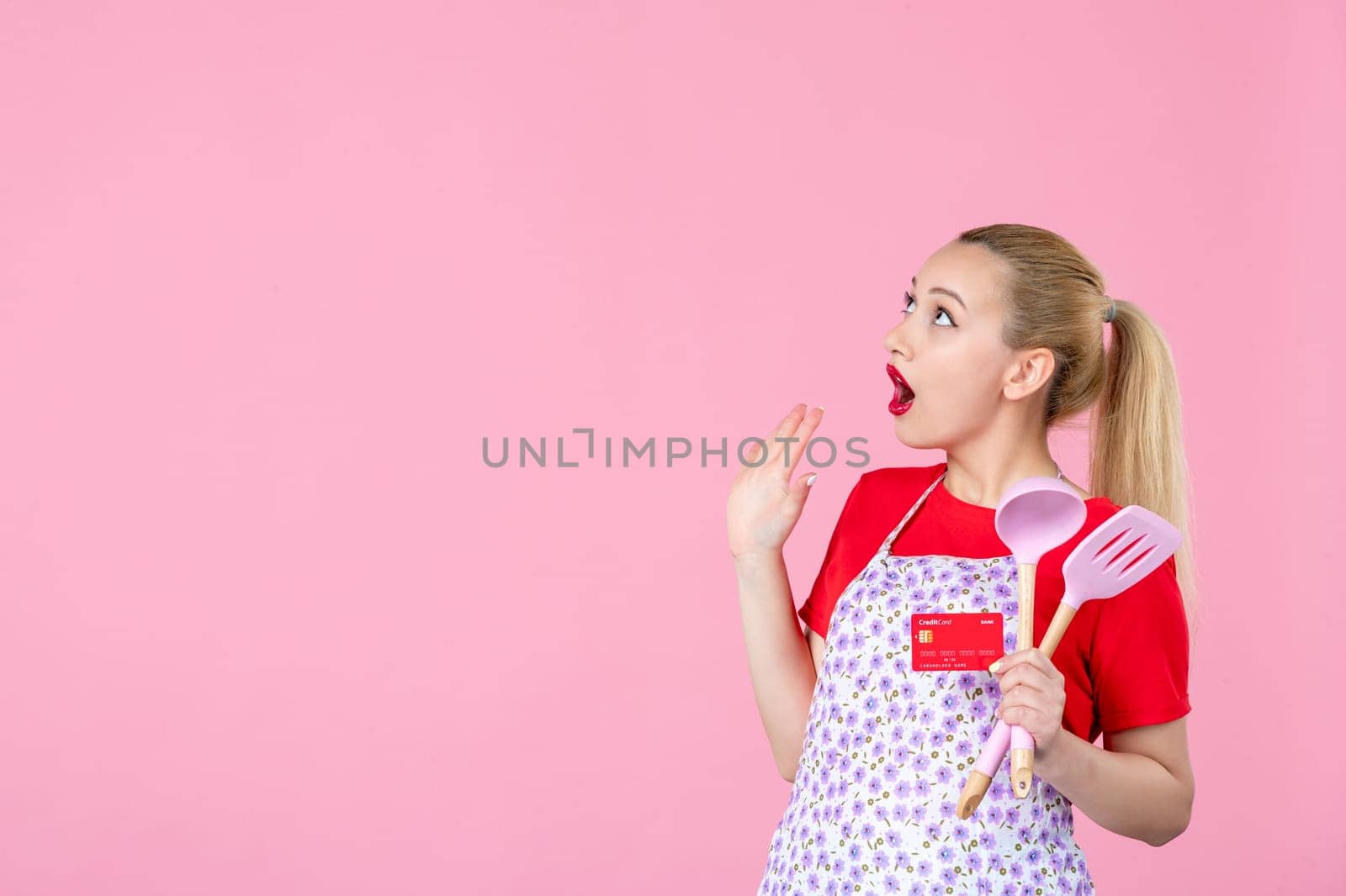 front view young housewife in cape holding spoons and red bank card on pink background occupation uniform profession job horizontal wife money duty workers