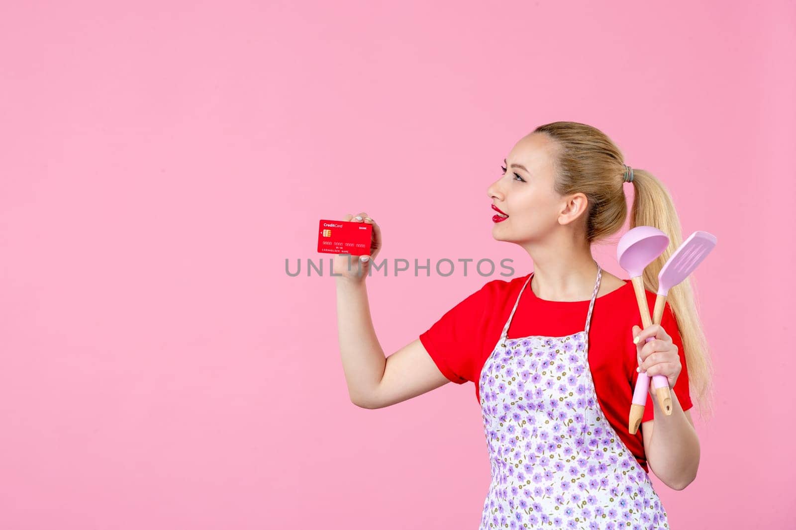 front view young housewife in cape holding spoons and red bank card on pink background profession money occupation duty job horizontal wife