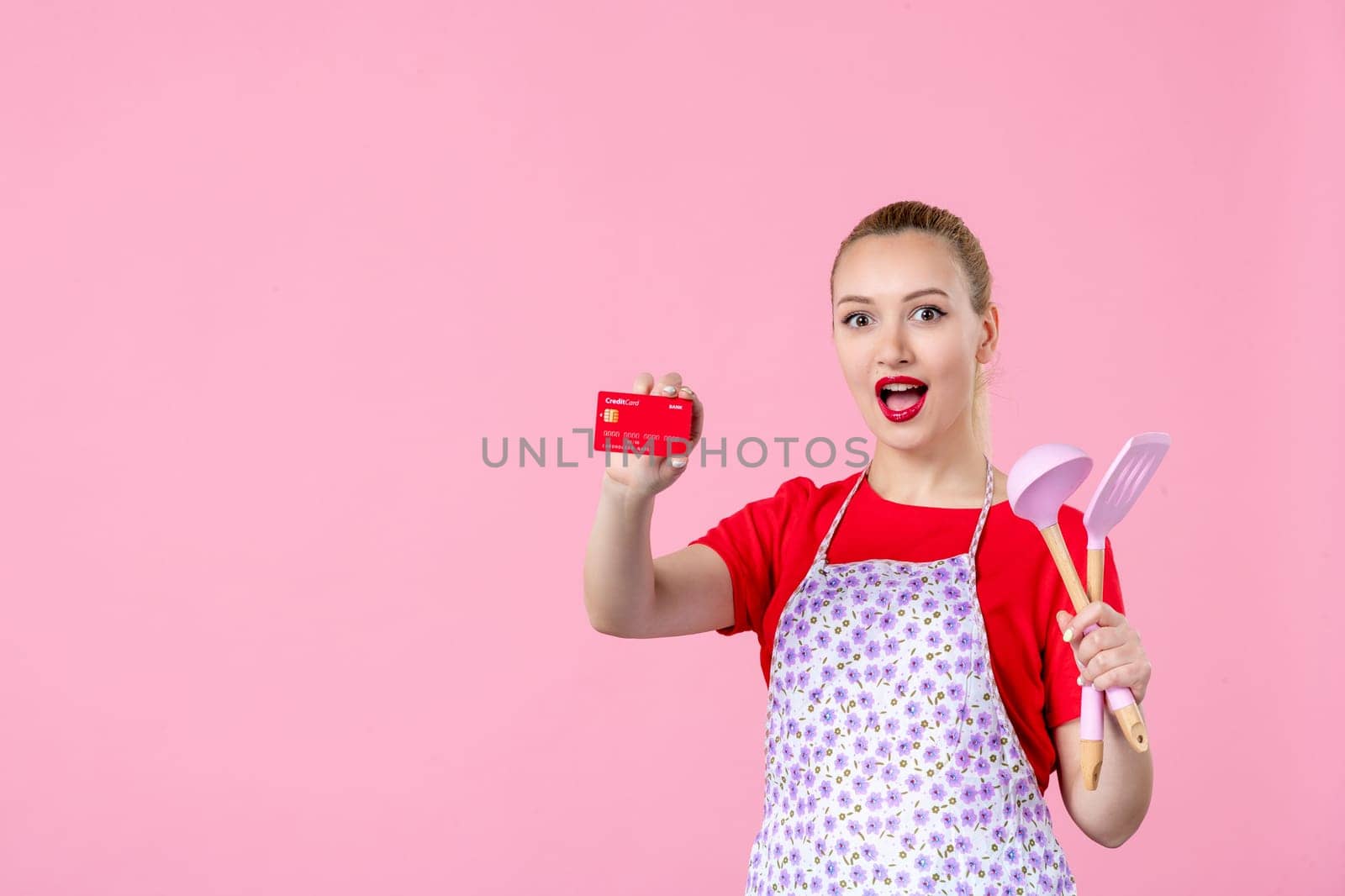 front view young housewife in cape holding spoons and red bank card on pink background profession money occupation duty uniform job cutlery horizontal wife