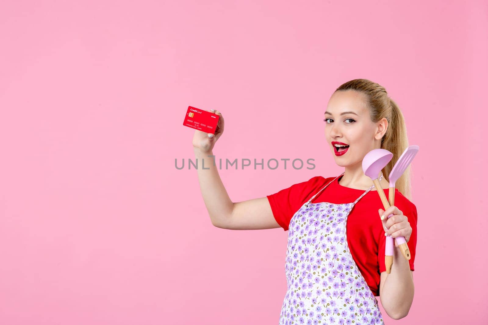 front view young housewife in cape holding spoons and red bank card on pink background profession money occupation duty uniform job cutlery worker horizontal