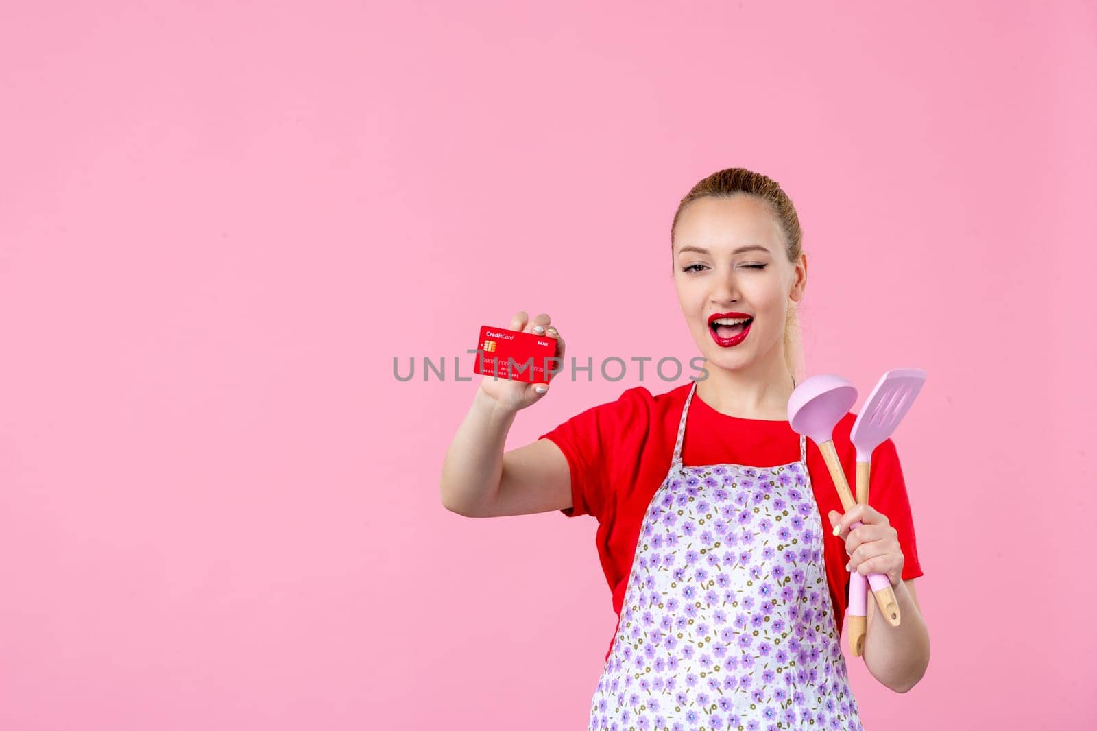 front view young housewife in cape holding spoons and red bank card on pink background profession money occupation duty uniform job cutlery worker horizontal wife