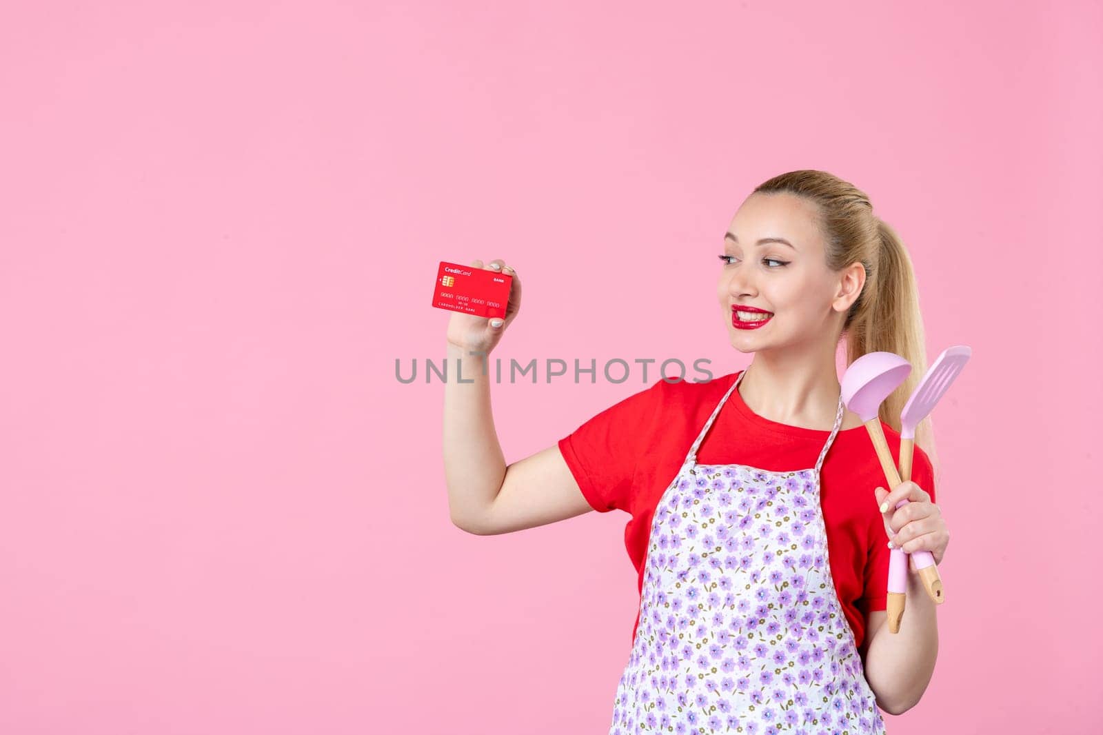 front view young housewife in cape holding spoons and red bank card on pink background profession money occupation duty uniform job cutlery worker wife by Kamran
